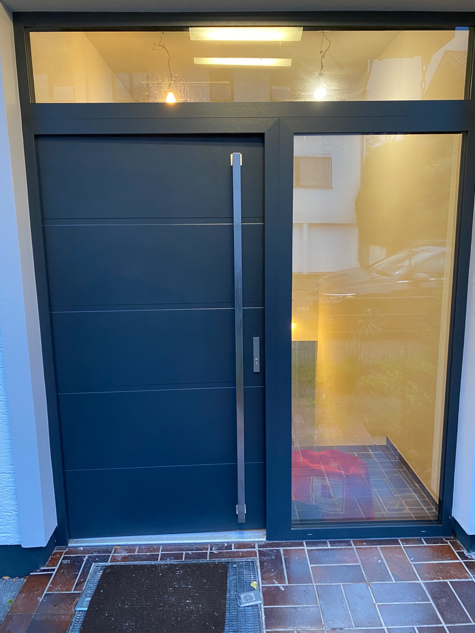 Nowoczesne, antracytowe drzwi wejściowe z długim, pionowym uchwytem ze stali nierdzewnej i szklanym panelem bocznym, widoczne płytki chodnikowe i wycieraczka przed wejściem. W szklanym panelu...