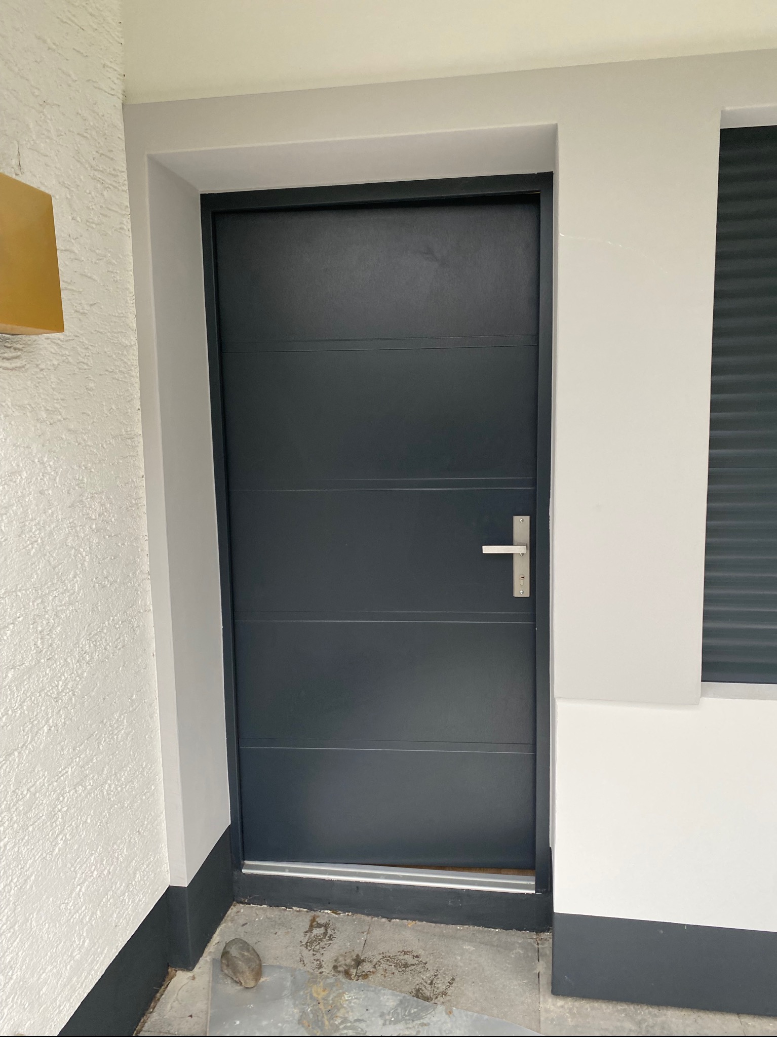 Nowoczesne, grafitowe drzwi wejściowe z poziomymi panelami i srebrną klamką, osadzone w białym murze z teksturą, fragment okna z żaluzjami w tle.