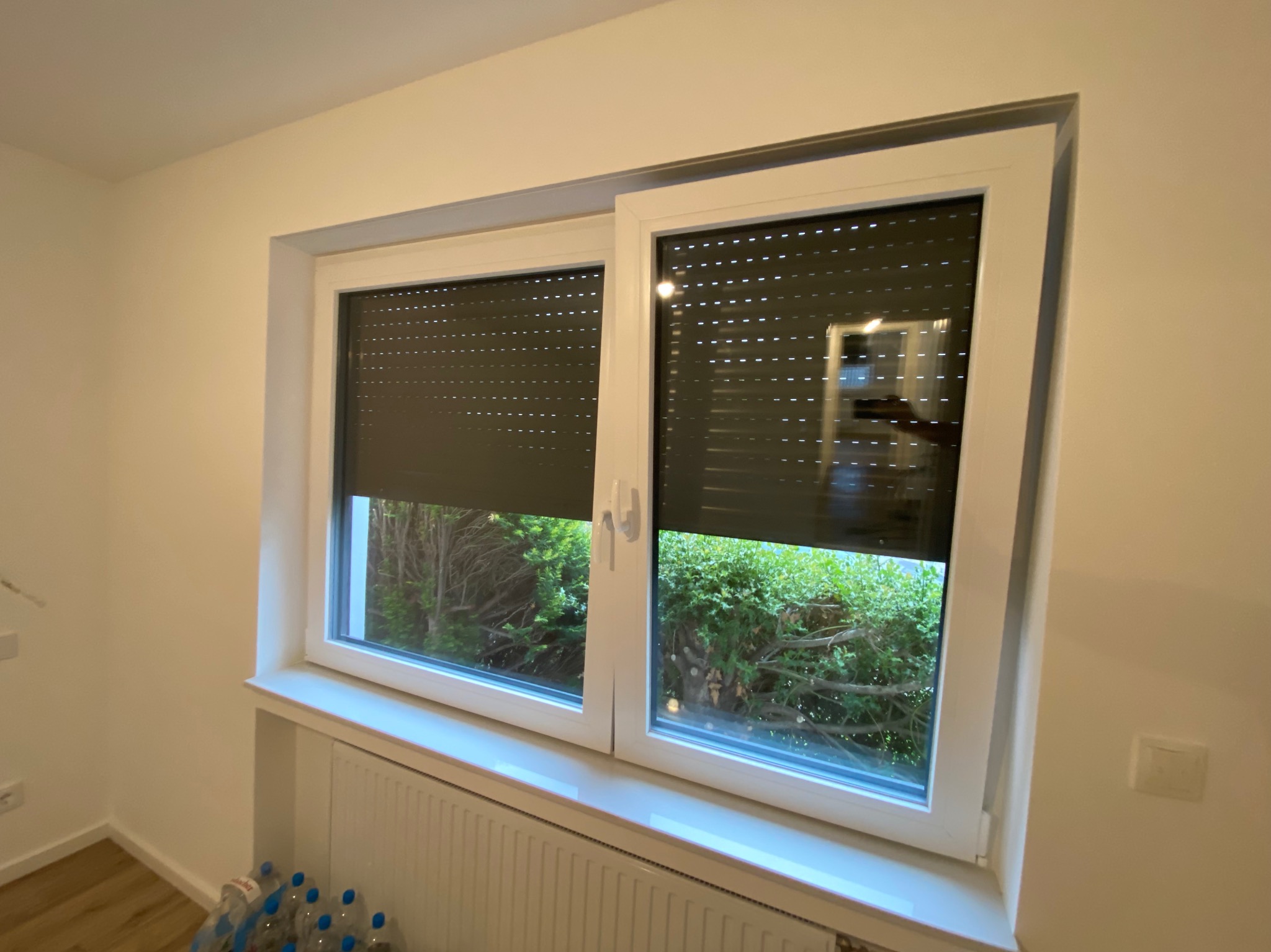 Okna PCV z roletami zewnętrznymi, częściowo opuszczonymi, widoczne z wnętrza pomieszczenia w Kędzierzynie-Koźlu, na parapecie grzejnik, za oknem zieleń ogrodu.