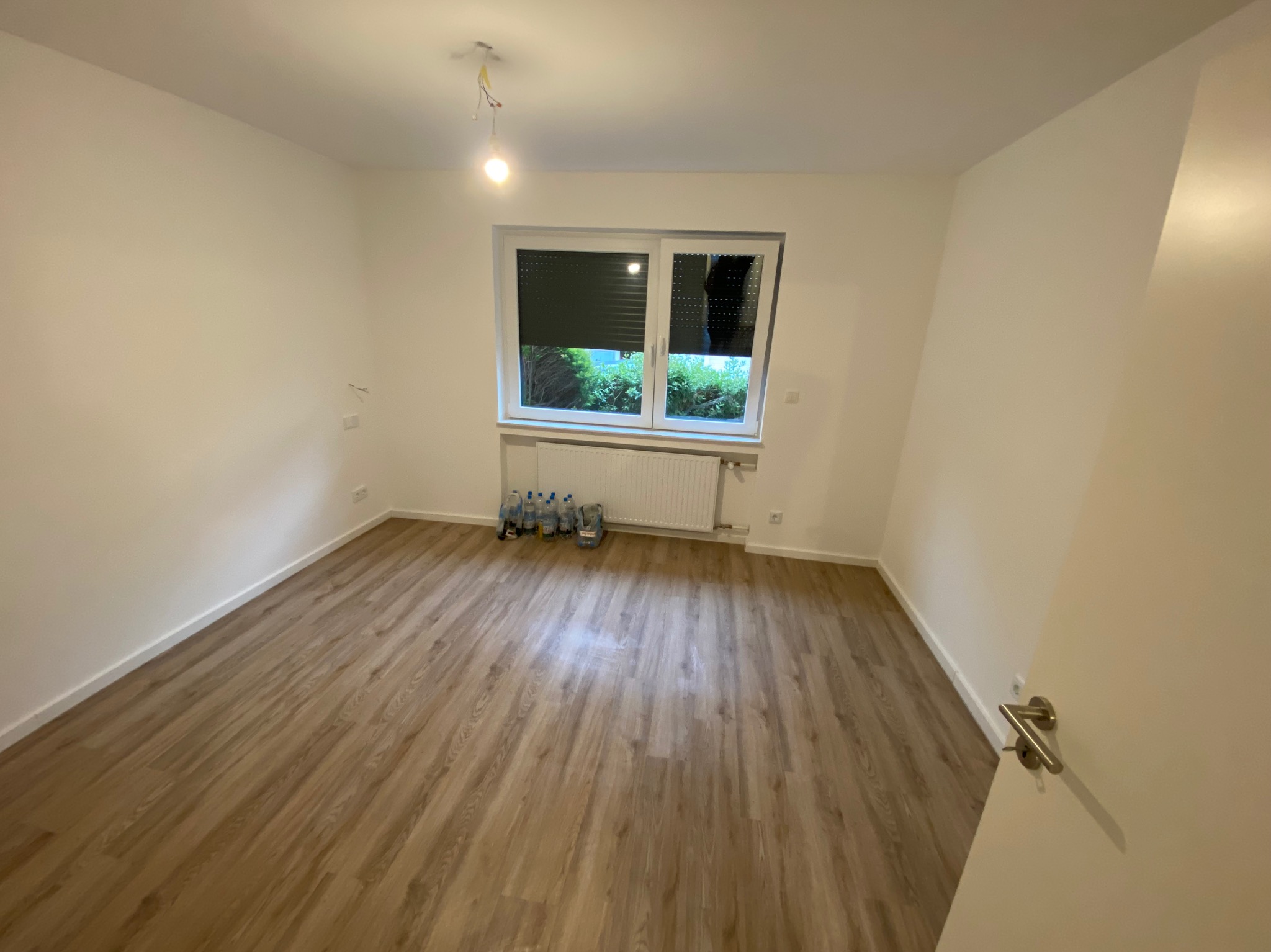 Pusty pokój z panelami podłogowymi, białymi ścianami, grzejnikiem pod oknem z częściowo zasłoniętymi roletami i wiszącą żarówką bez klosza. Na podłodze, pod oknem, stoi kilka butelek z wodą i inne...