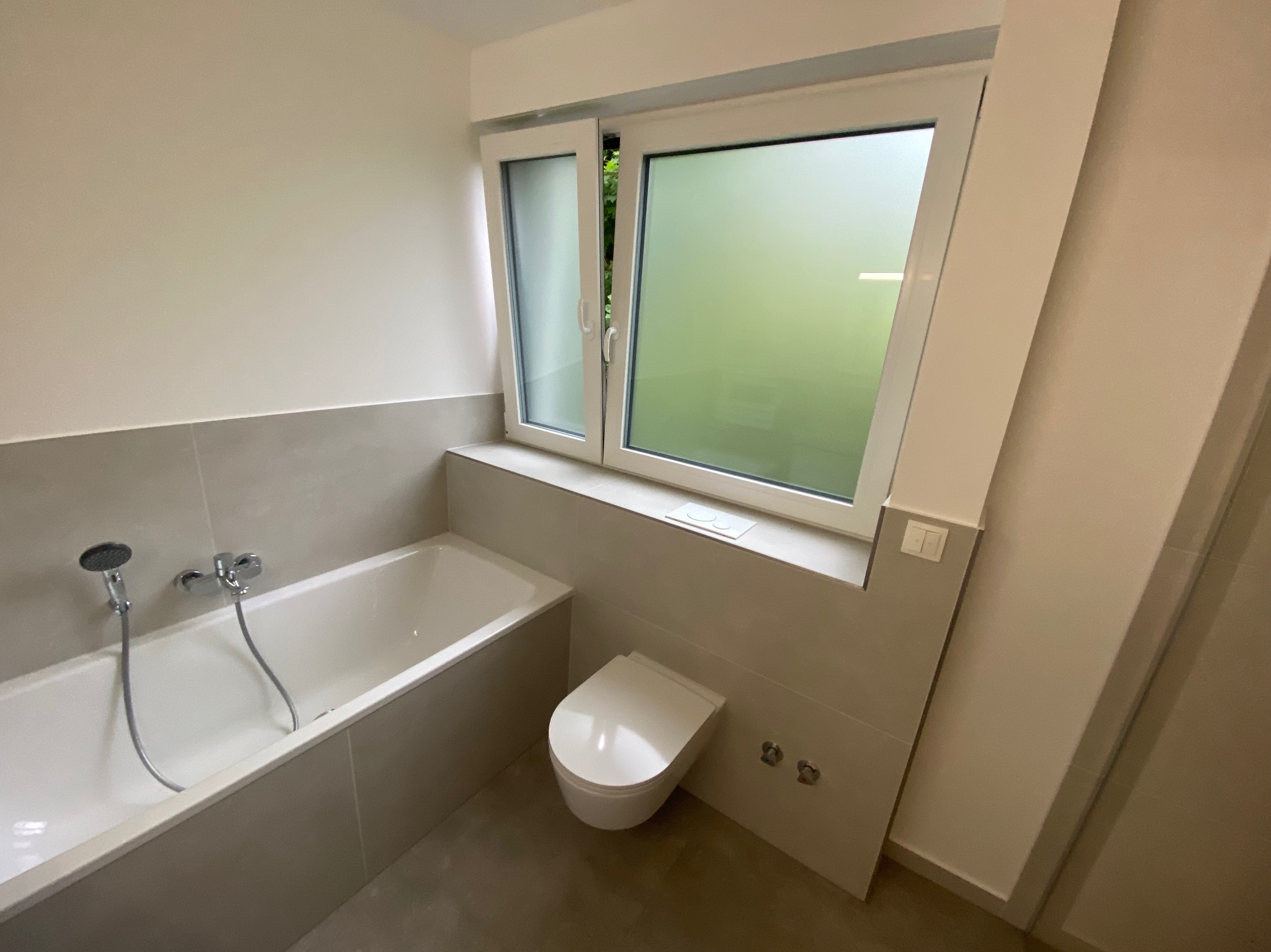 Biała wanna, podwieszana toaleta i okno z mleczną szybą w wykończonej łazience z szarymi płytkami.