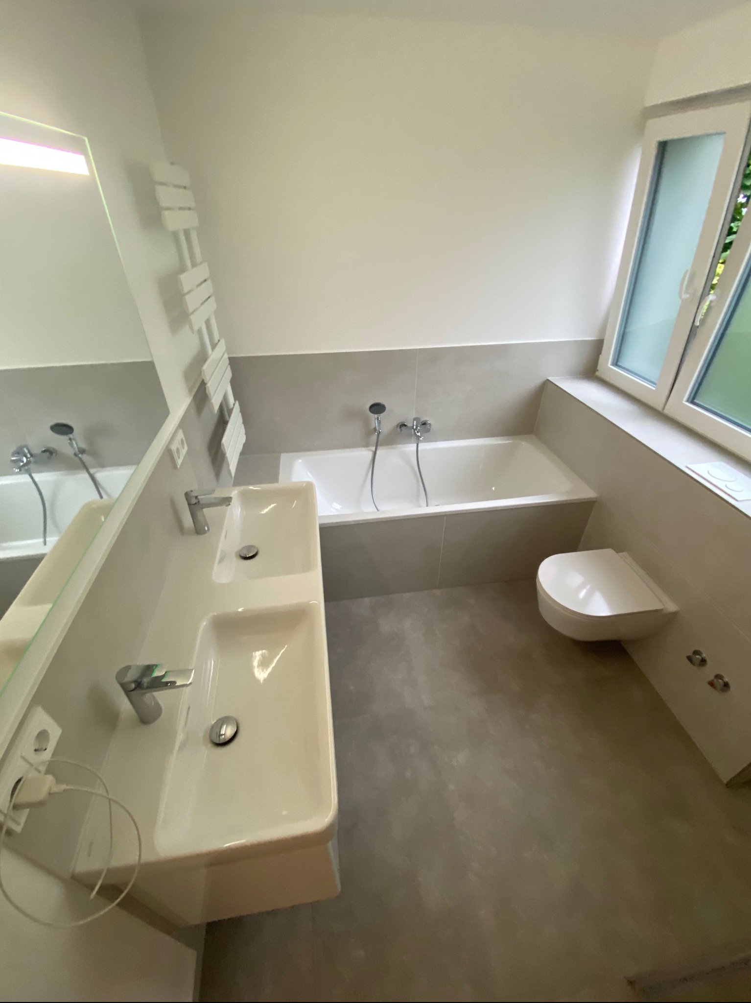 Nowoczesna łazienka z dwiema umywalkami, wanną i wiszącą toaletą, utrzymana w szaro-białej kolorystyce, z grzejnikiem drabinkowym i dużym oknem.