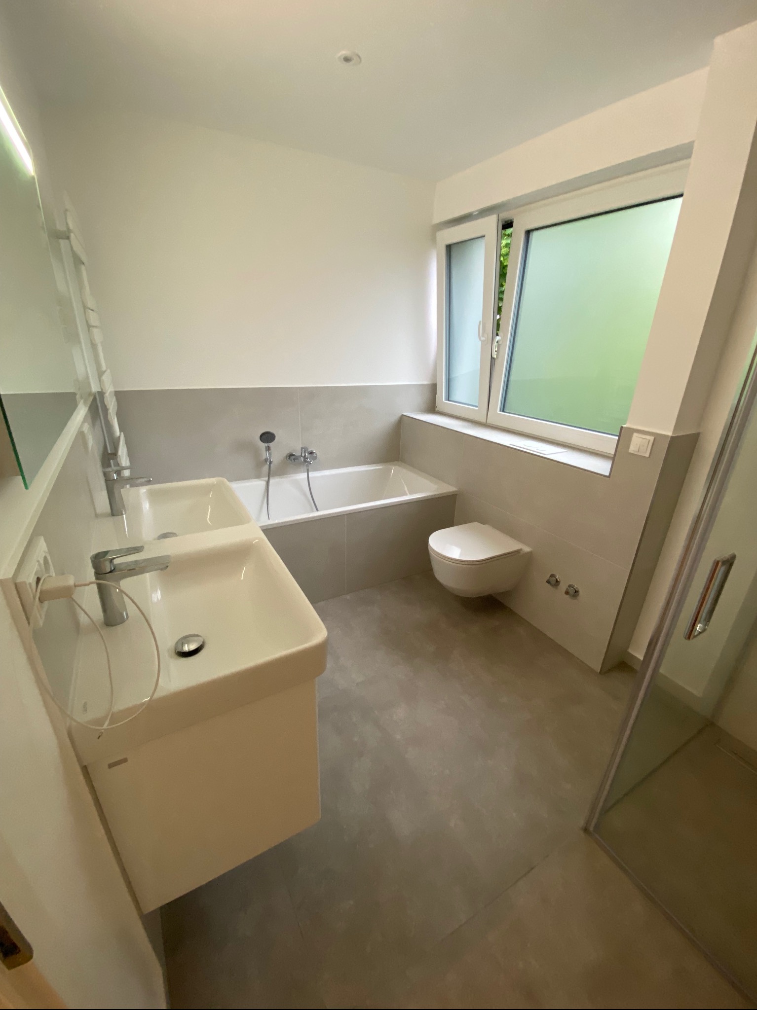 Nowoczesna łazienka z dwiema umywalkami nablatowymi, wanną, toaletą podwieszaną i kabiną prysznicową z widokiem na zieleń za oknem.