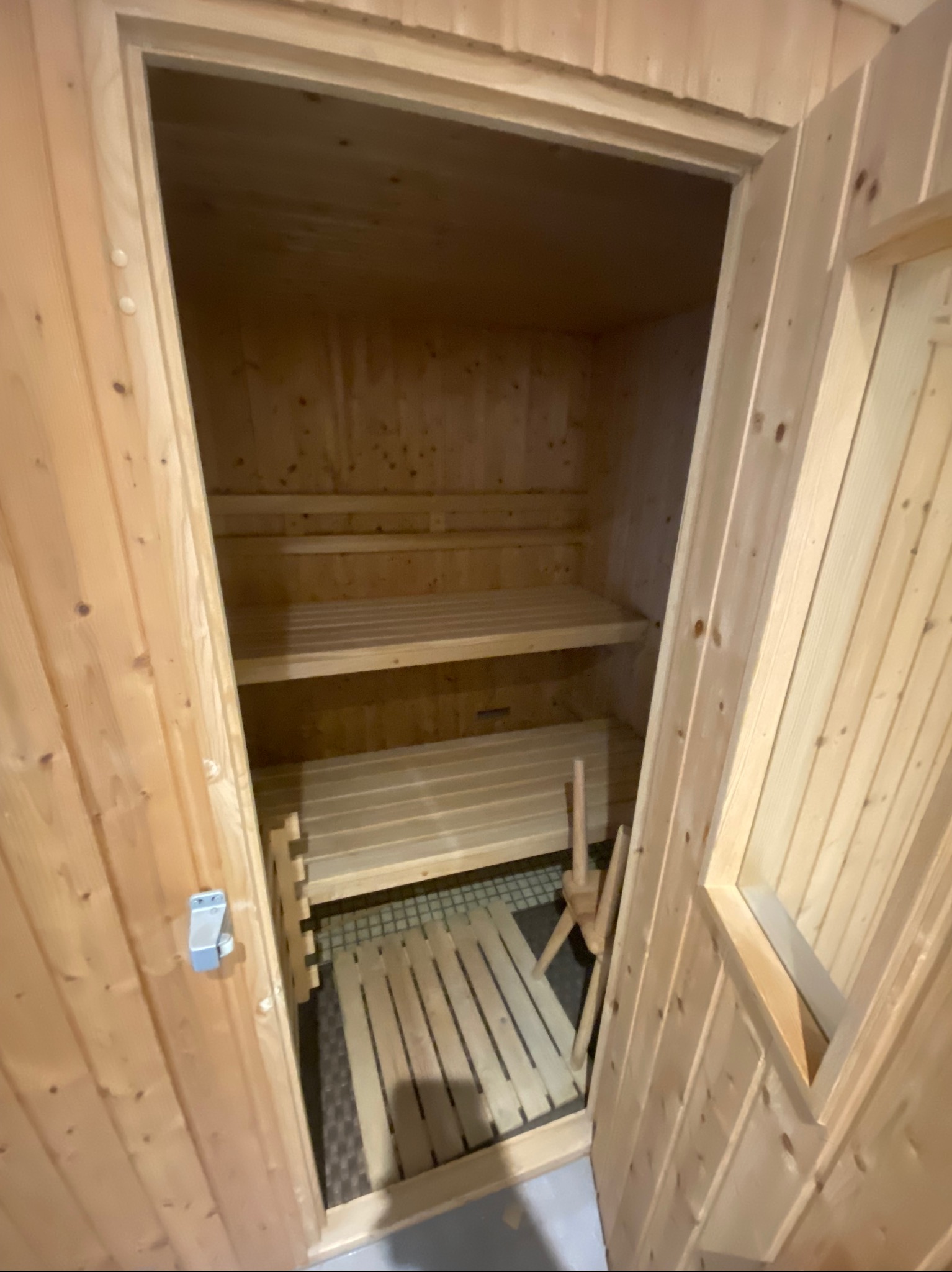 Wnętrze nowej sauny z jasnego drewna, widoczne półki i siedziska, fragment pieca. Drzwi otwarte, z widocznym metalowym zamkiem.