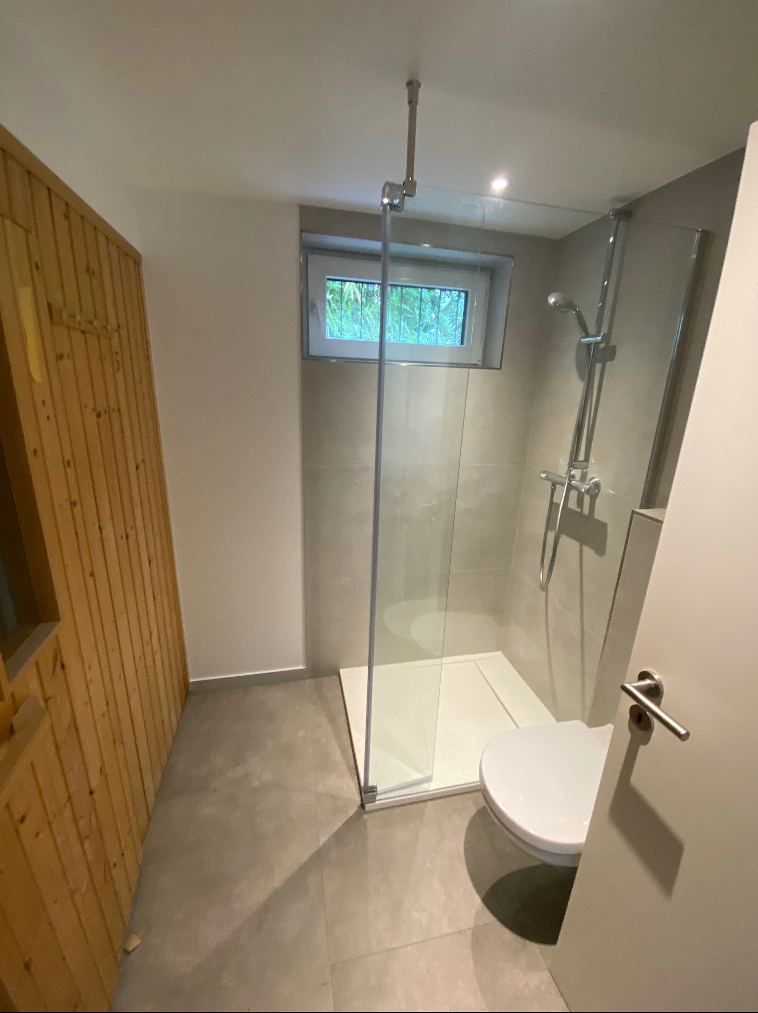 Nowoczesna łazienka z kabiną prysznicową typu walk-in, wykończona szarymi płytkami i drewnianą boazerią, z widokiem na zieleń za oknem.