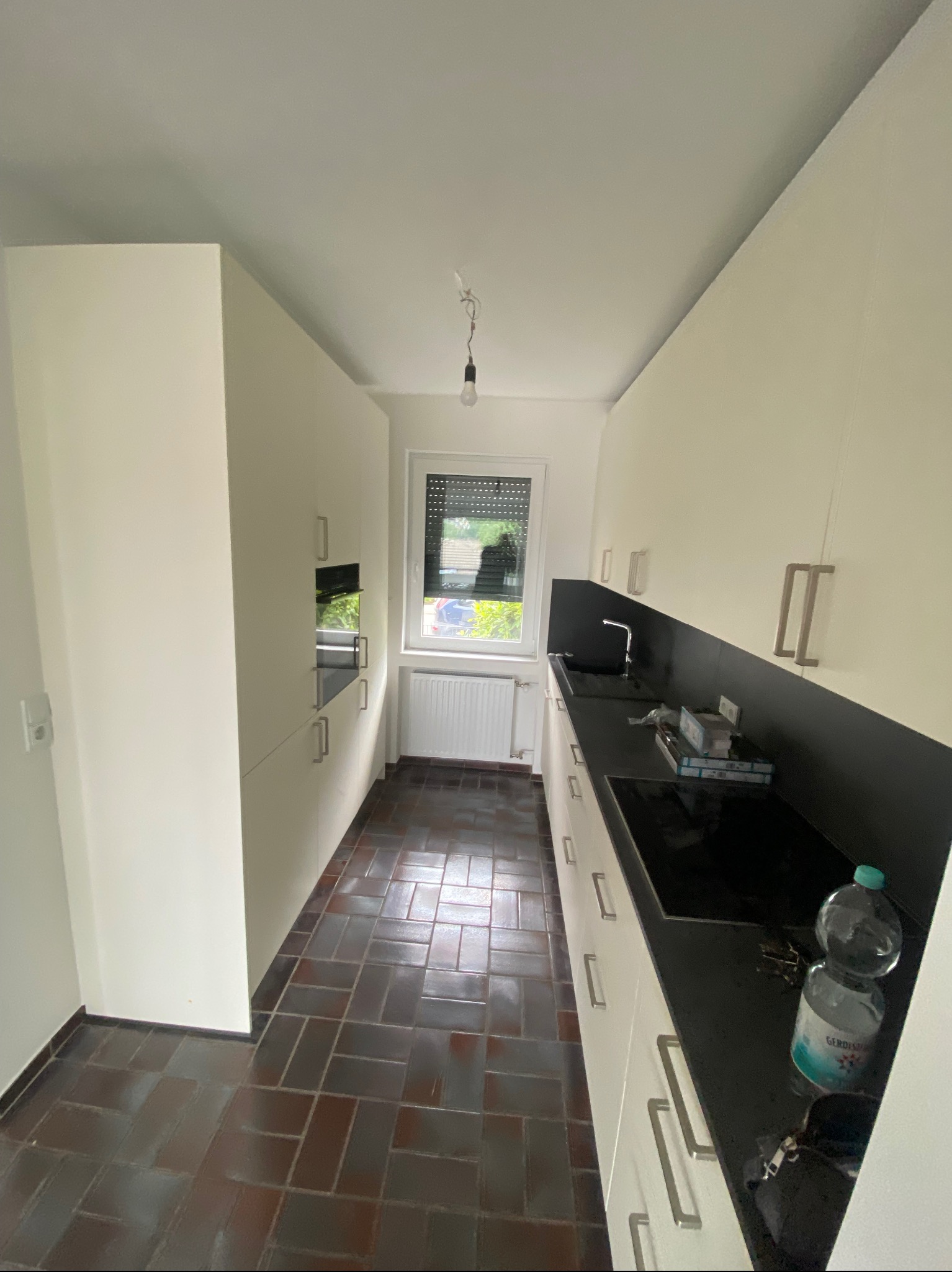 Wnętrze kuchni z brązową podłogą z płytek, białymi szafkami z długimi, srebrnymi uchwytami i czarnym blatem, widok z perspektywy wejścia.