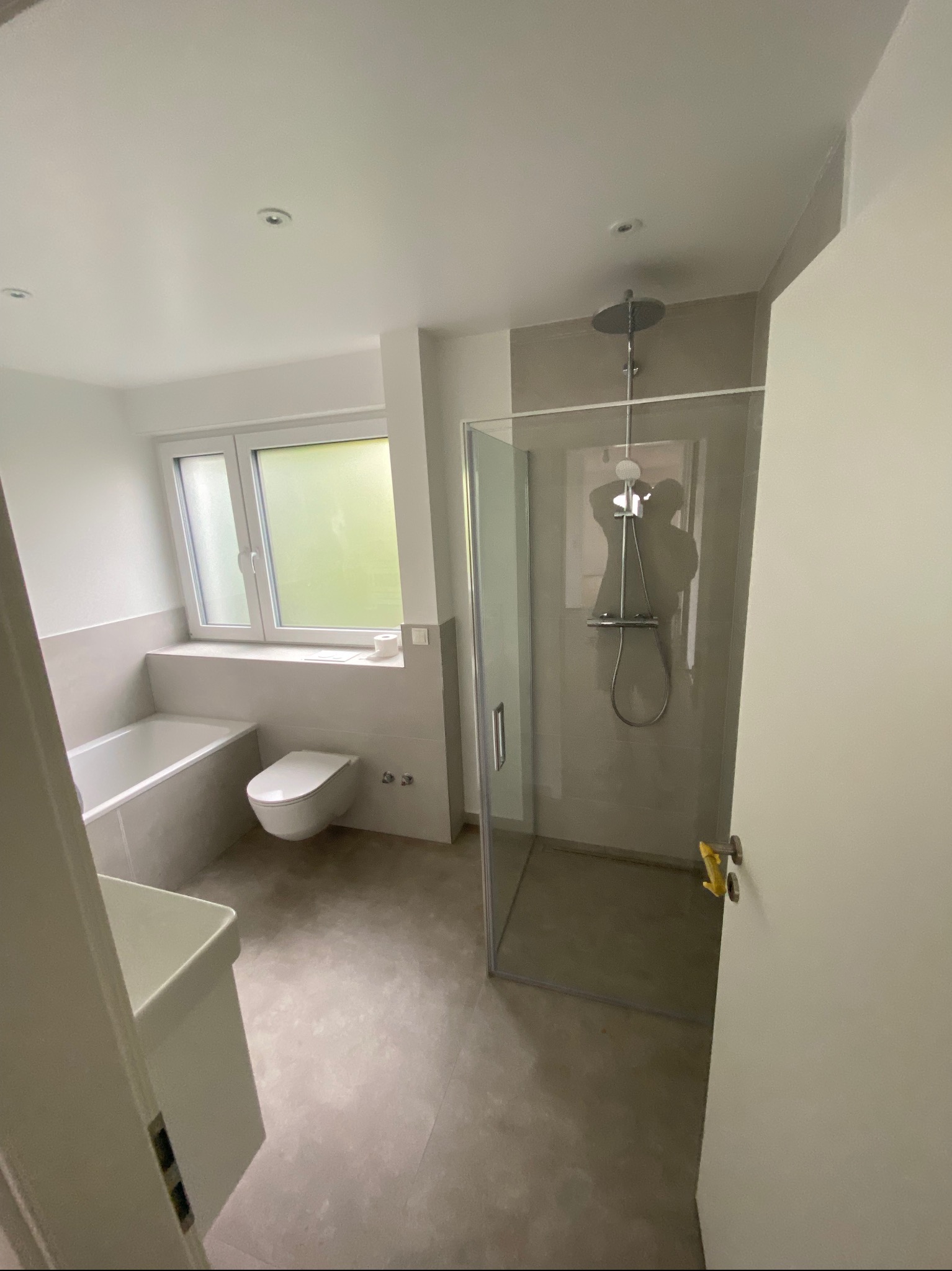 Nowoczesna łazienka z kabiną prysznicową typu walk-in, umywalką, wanną i toaletą, wykończona w odcieniach szarości i bieli, z oknem zapewniającym naturalne światło.