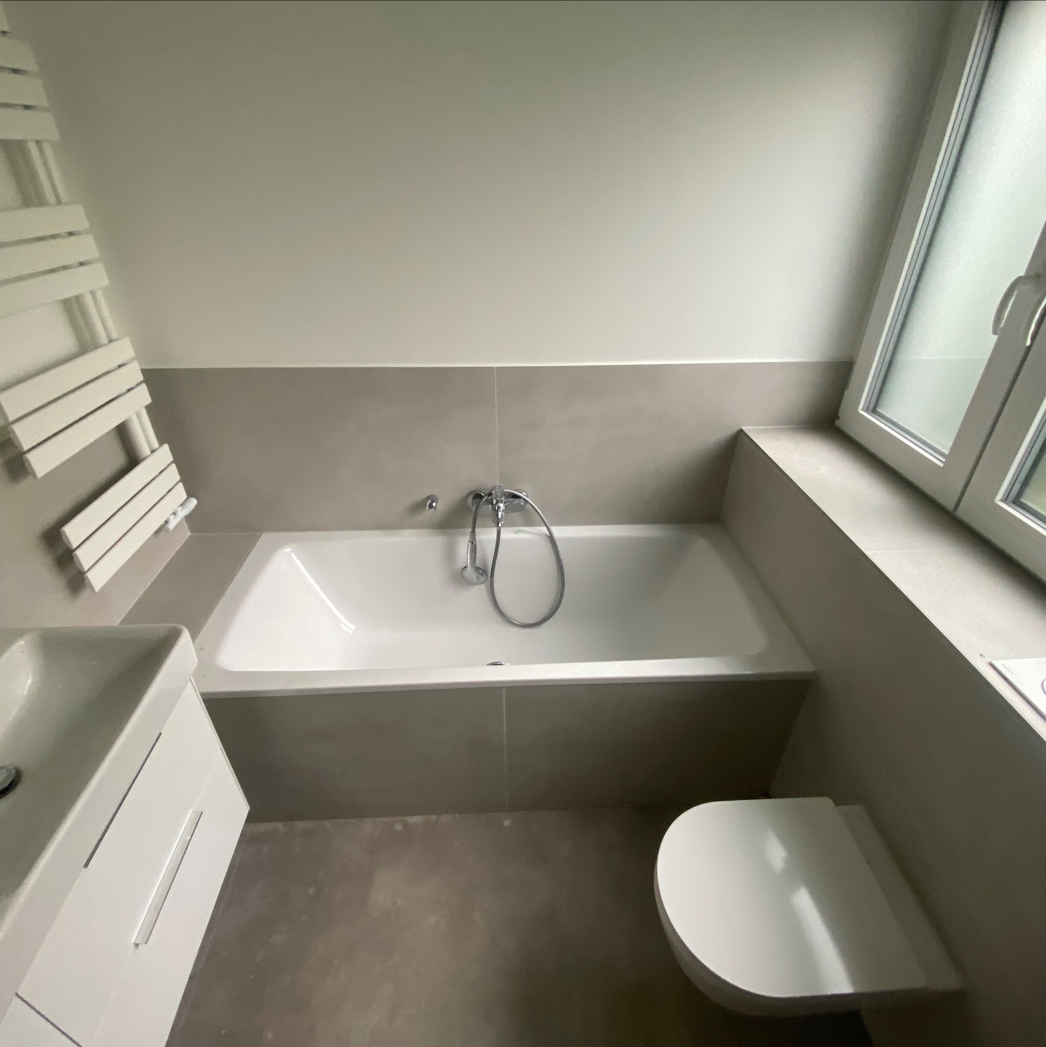 Nowoczesna łazienka w neutralnych barwach z białą armaturą: wanna z baterią, umywalka z szafką i toaleta. Na ścianie grzejnik drabinkowy.