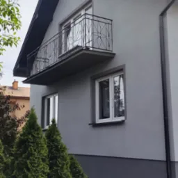 Szara elewacja domu jednorodzinnego z balkonem o dekoracyjnej, metalowej balustradzie, widoczna z poziomu kostki brukowej, z fragmentem rynny i zielonymi tujami wzdłuż ściany.