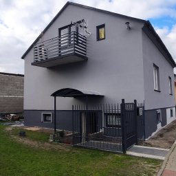 Damian Sadowski - Szary dom jednorodzinny z balkonem, świeżo po malowaniu, widoczna podmurówka w innym odcieniu szarości, teren wokół częściowo uporządkowany, w tle kościelna wieża.