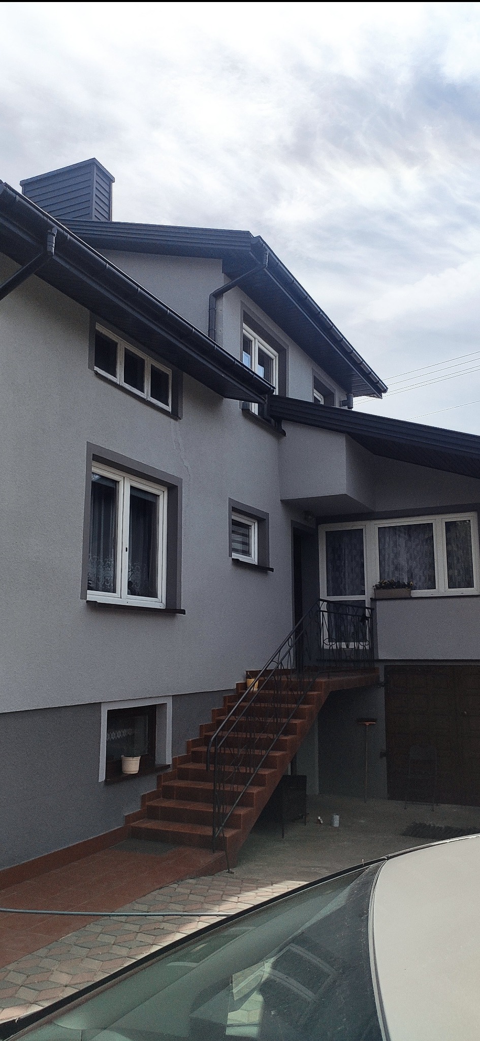 Dwukondygnacyjny dom z szarą elewacją, brązowymi schodami wejściowymi i ciemnymi obramowaniami okien oraz dachu, widoczny zza szyby samochodu.