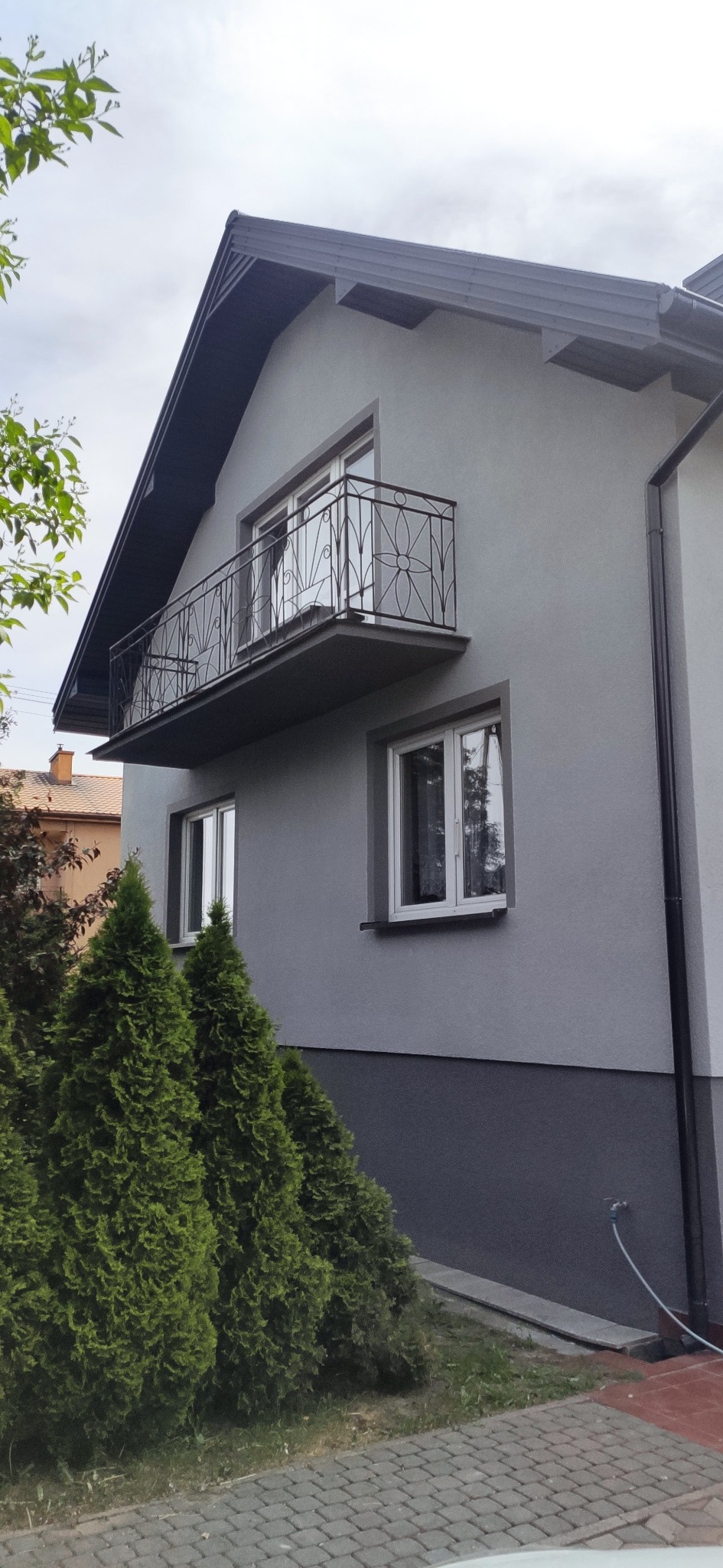 Szara elewacja domu jednorodzinnego z balkonem o dekoracyjnej, metalowej balustradzie, widoczna z poziomu kostki brukowej, z fragmentem rynny i zielonymi tujami wzdłuż ściany.