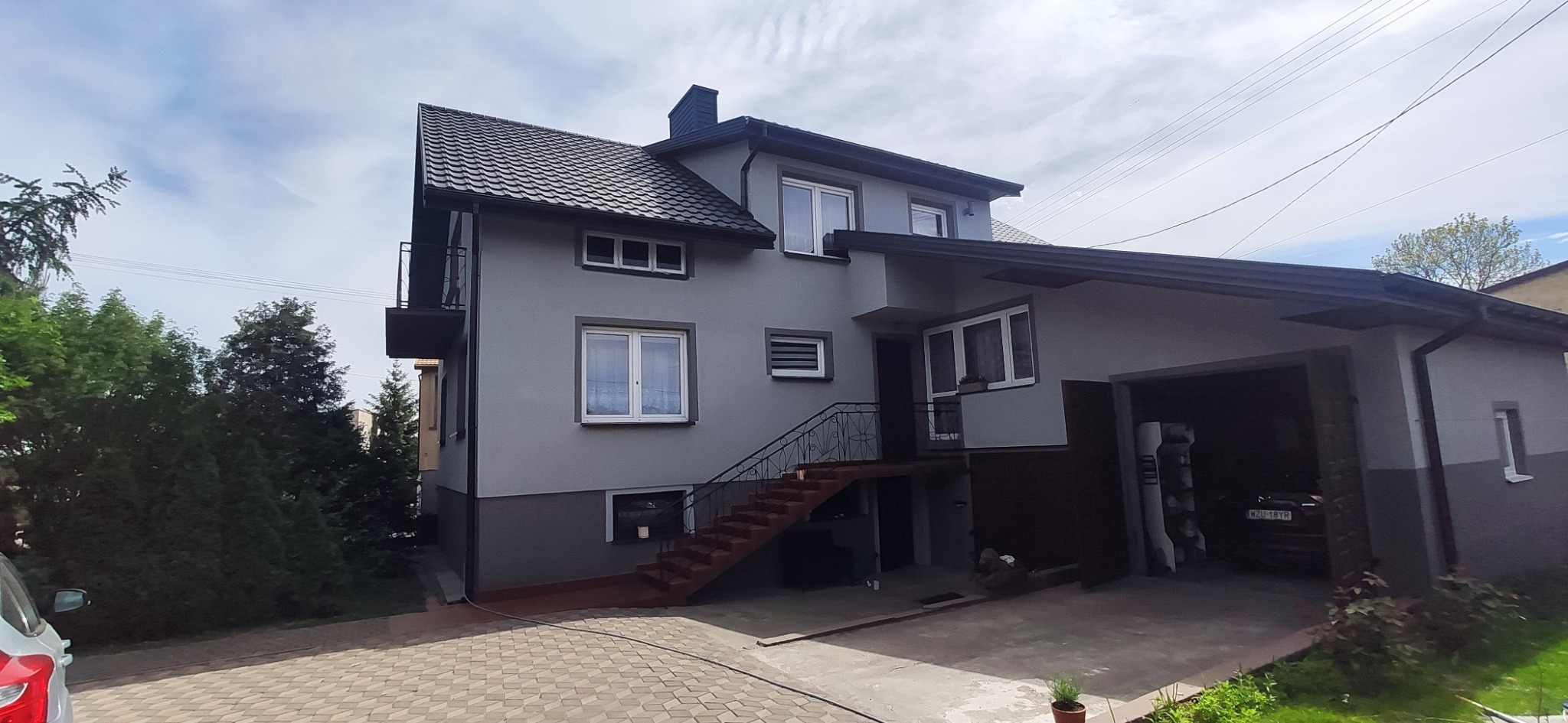 Dwukondygnacyjny dom z szarą elewacją, czarnym dachem i schodami wejściowymi z metalową balustradą, widok z zewnątrz, w tle błękitne niebo z chmurami.