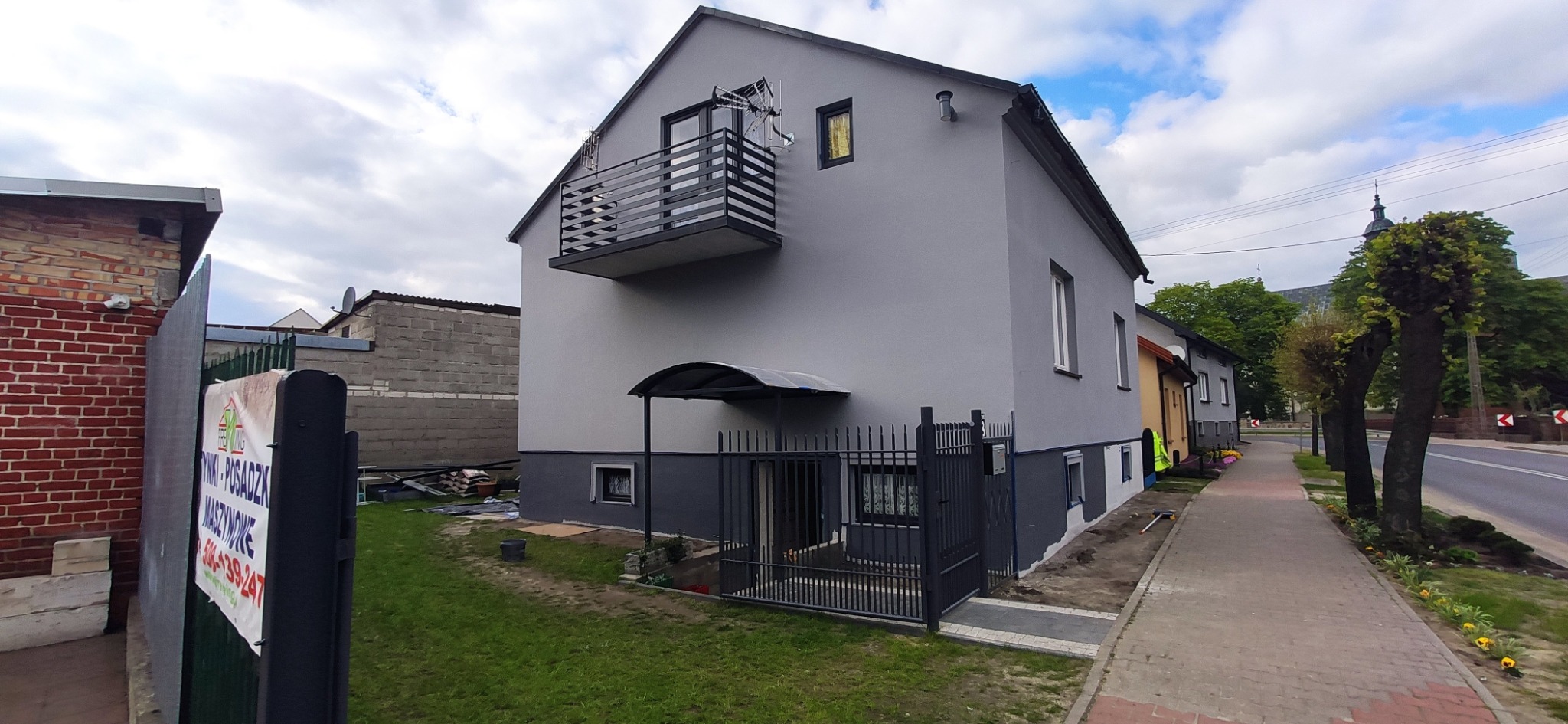 Szary dom jednorodzinny z balkonem, świeżo po malowaniu, widoczna podmurówka w innym odcieniu szarości, teren wokół częściowo uporządkowany, w tle kościelna wieża.