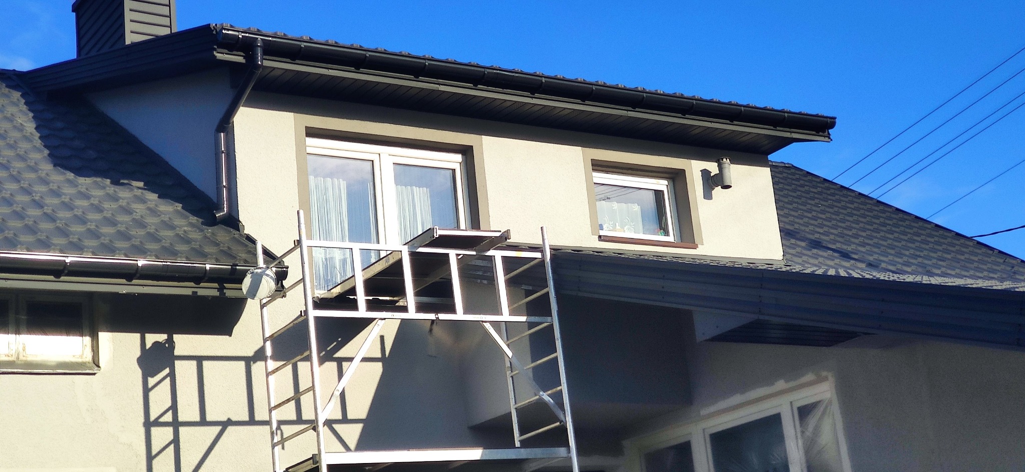 Elewacja domu w trakcie prac wykończeniowych z użyciem rusztowania, widoczne okna zabezpieczone folią malarską oraz fragment dachu z rynnami na tle błękitnego nieba.