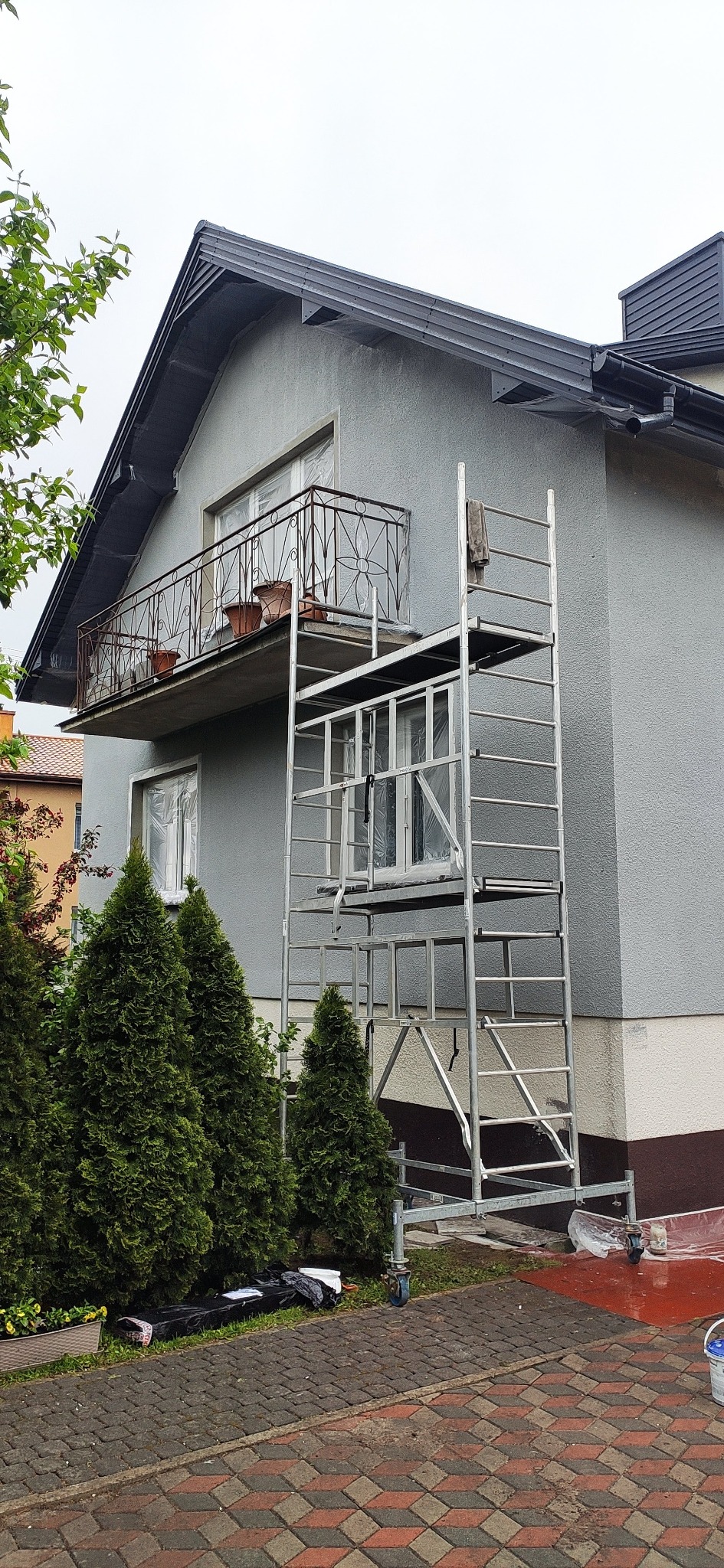 Szara elewacja domu jednorodzinnego w trakcie malowania, z balkonem z ozdobną balustradą, osłoniętymi oknami i rozstawionym rusztowaniem, widok z poziomu kostki brukowej.