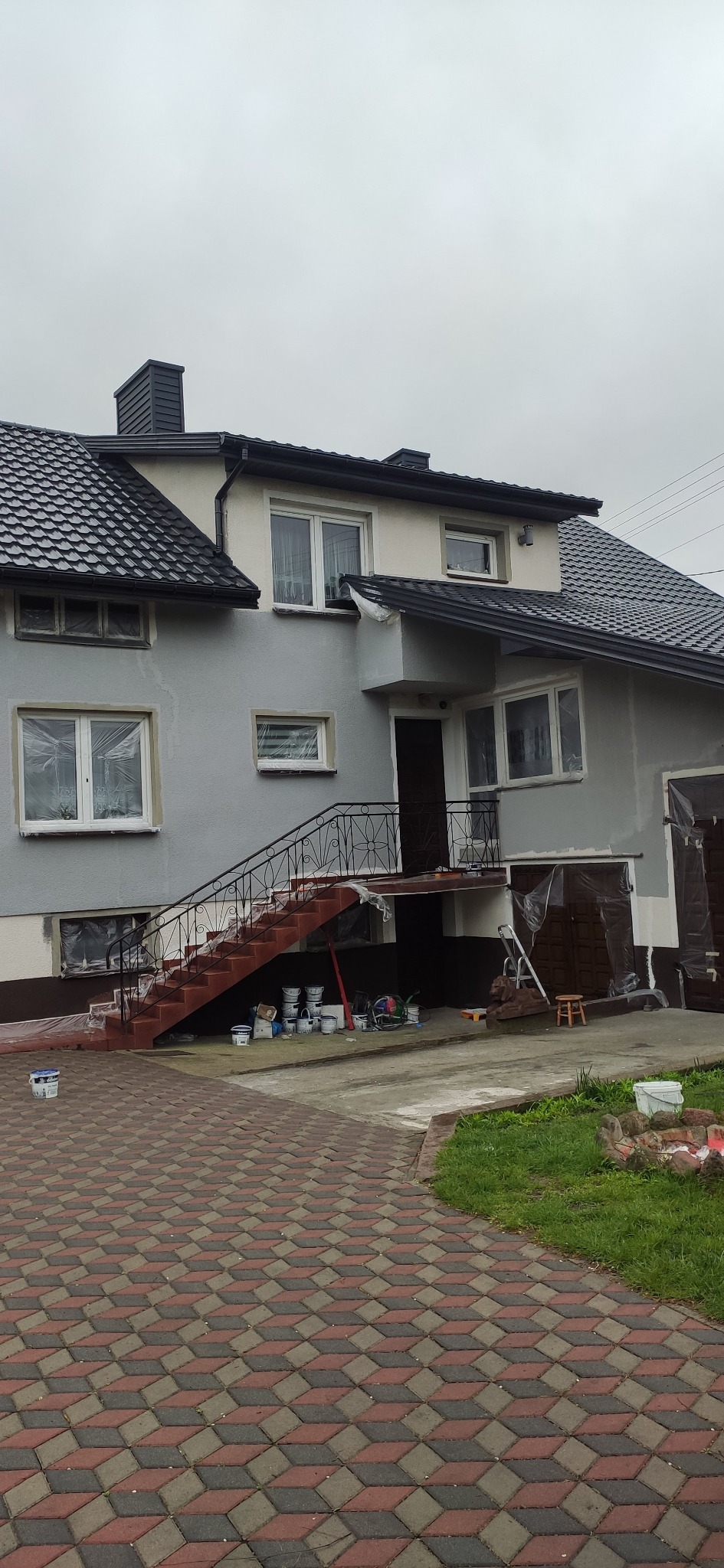 Dwukondygnacyjny dom z szarą elewacją w trakcie prac wykończeniowych, okna zabezpieczone folią, widoczne puszki z farbą u podnóża schodów prowadzących do wejścia, świeżo pomalowane schody...