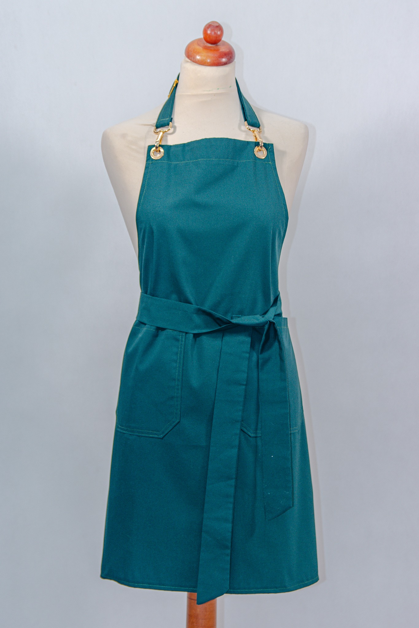 Turkusowy fartuch z kieszeniami i złotymi detalami, zawiązany w pasie, prezentowany na manekinie na jasnym tle. Widoczne szwy i wykończenie.