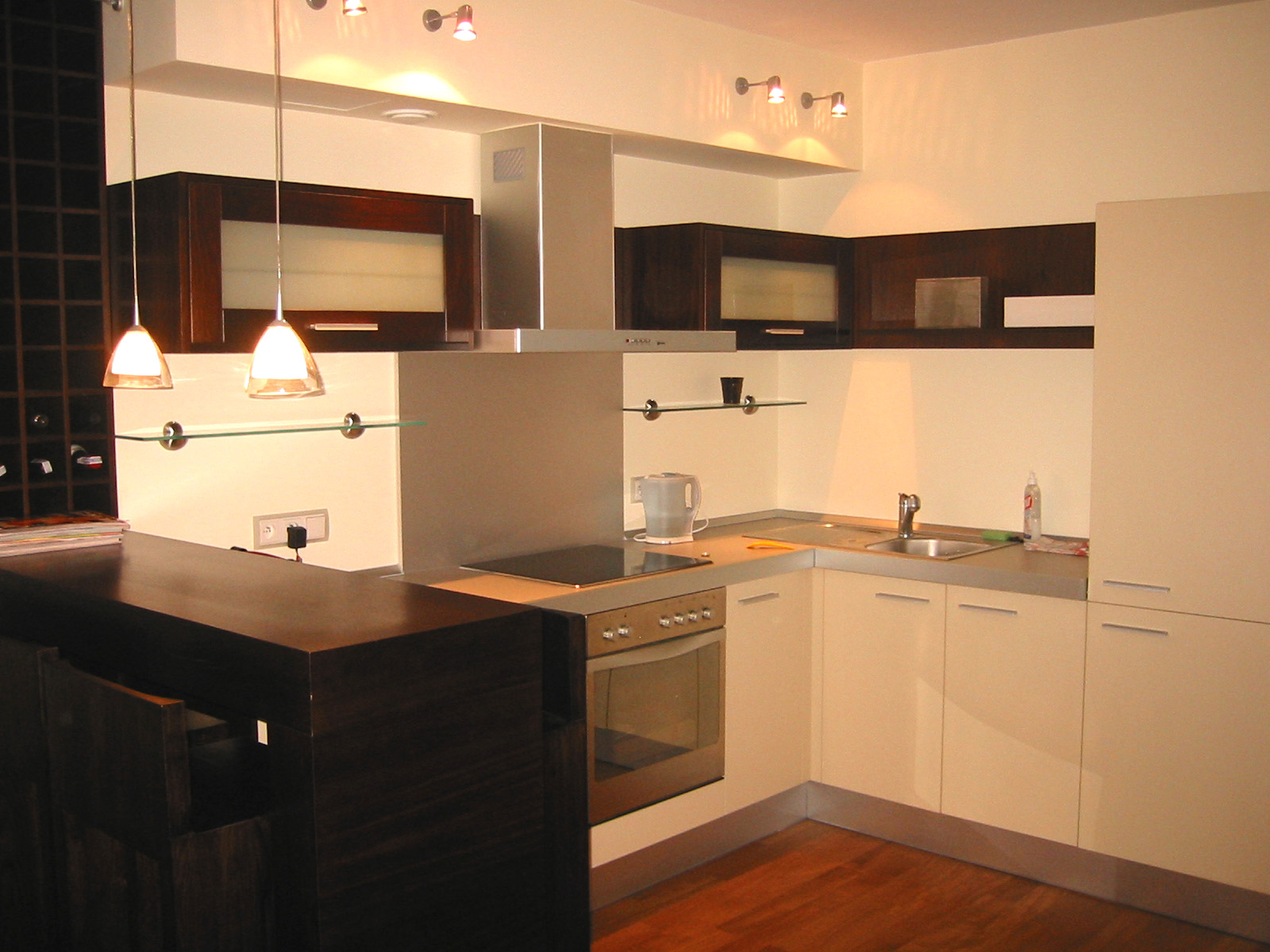 Nowoczesna kuchnia z drewnianym blatem barowym, białymi szafkami i ciemnymi, wiszącymi szafkami z podświetlanymi frontami, oświetlona wiszącymi lampami i halogenami.