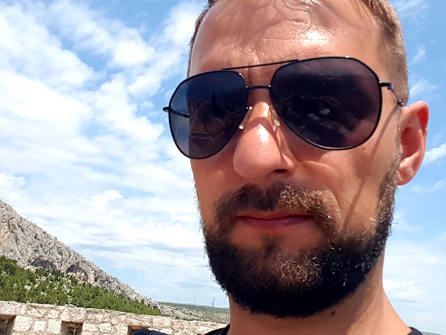 Portret mężczyzny w okularach przeciwsłonecznych na tle błękitnego nieba i skalistego krajobrazu. Widoczny fragment kamiennego muru.