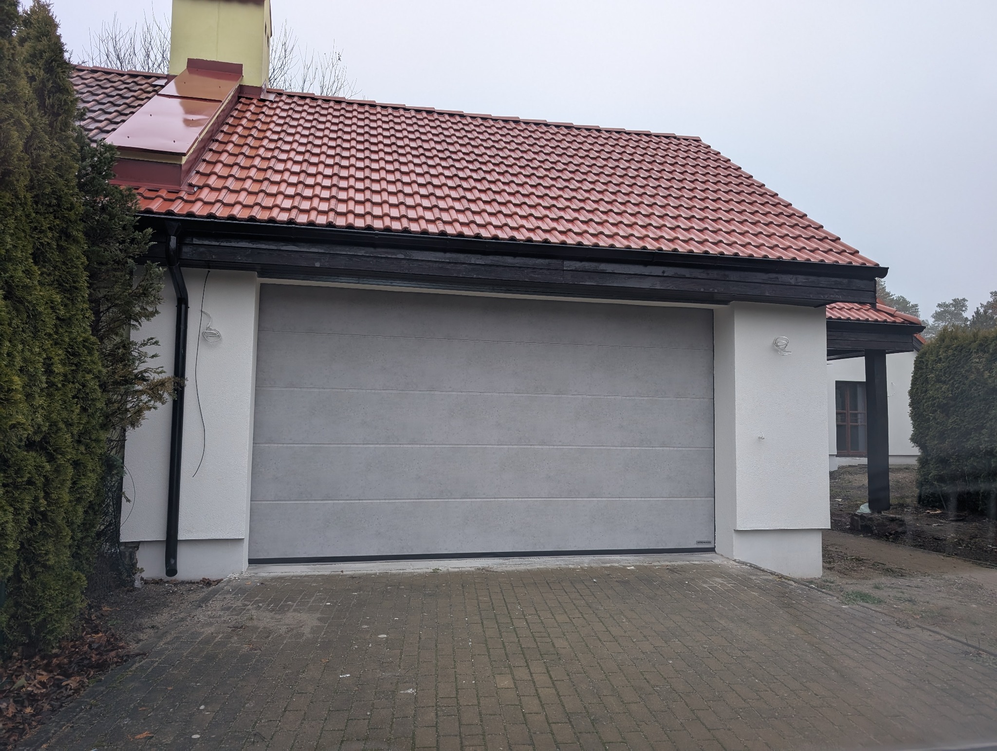 Szara segmentowa brama garażowa z poziomymi przetłoczeniami, zamontowana w białej elewacji domu z czerwoną dachówką, widok z zewnątrz na podjazd z kostki.