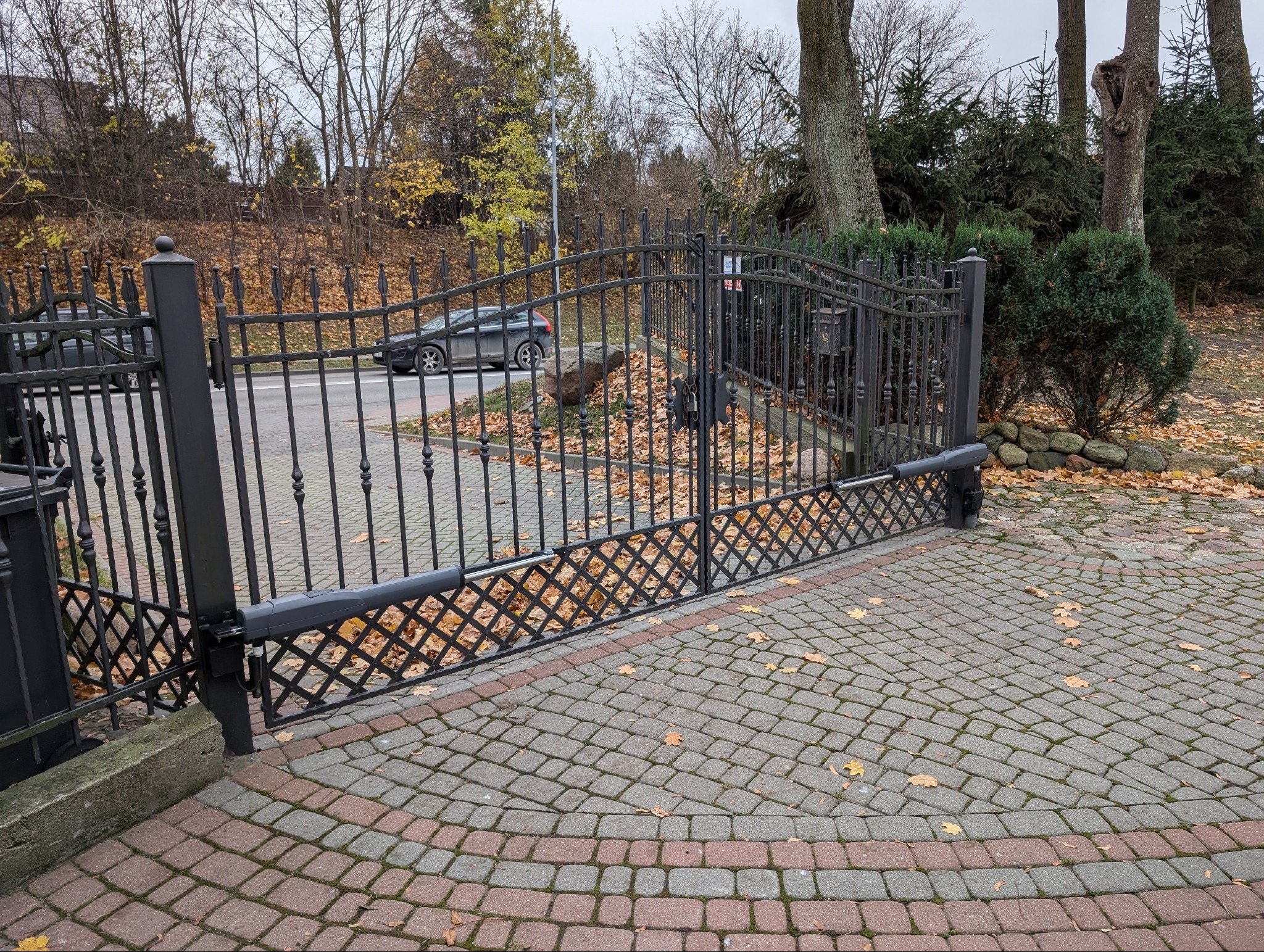 Automatyczna brama wjazdowa z czarnego metalu, widoczny mechanizm otwierający, kostka brukowa na podjeździe, jesienne liście.
