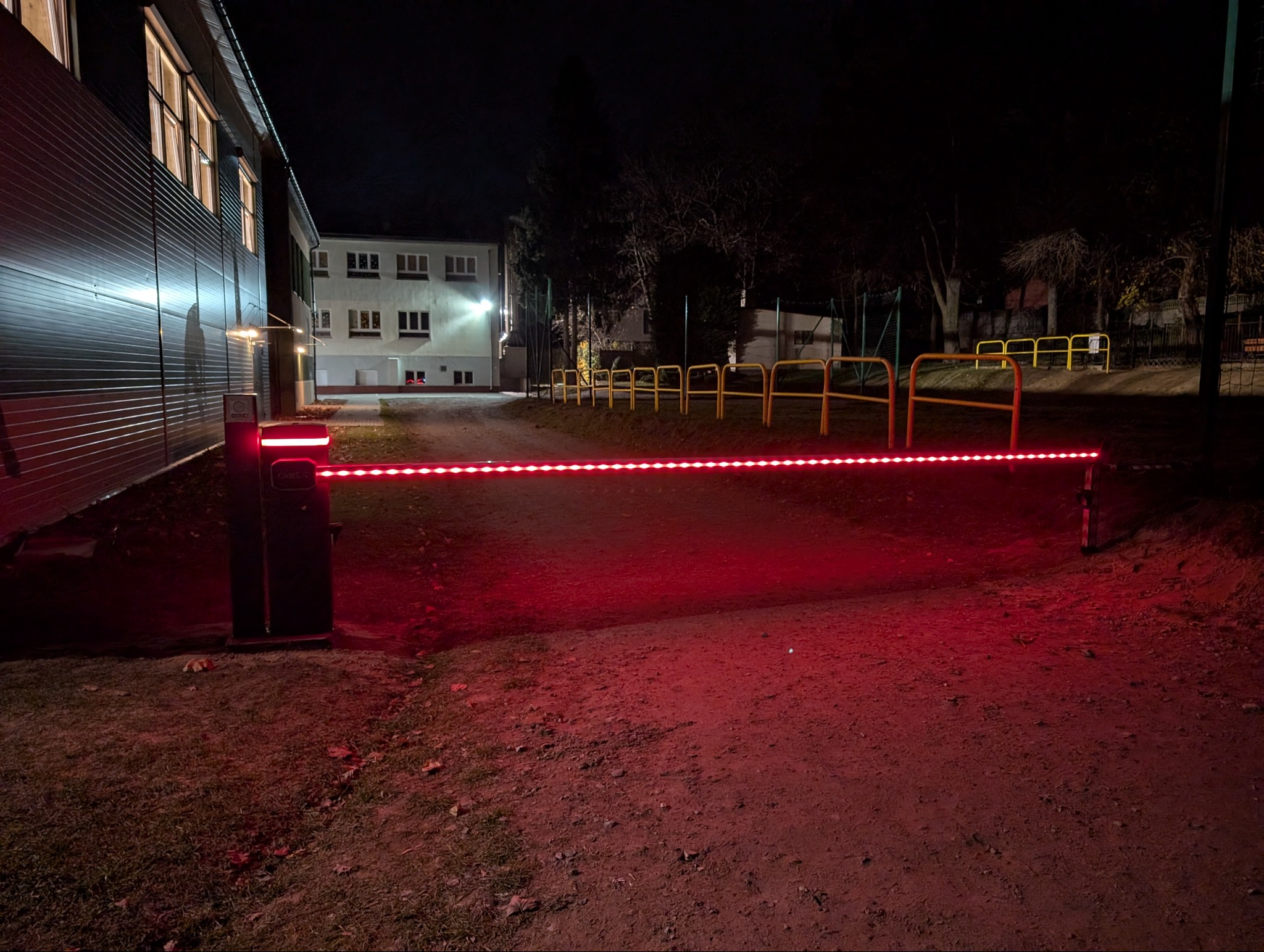 Szlaban automatyczny z czerwonym oświetleniem LED w pozycji zamkniętej, nocne ujęcie na tle budynków.