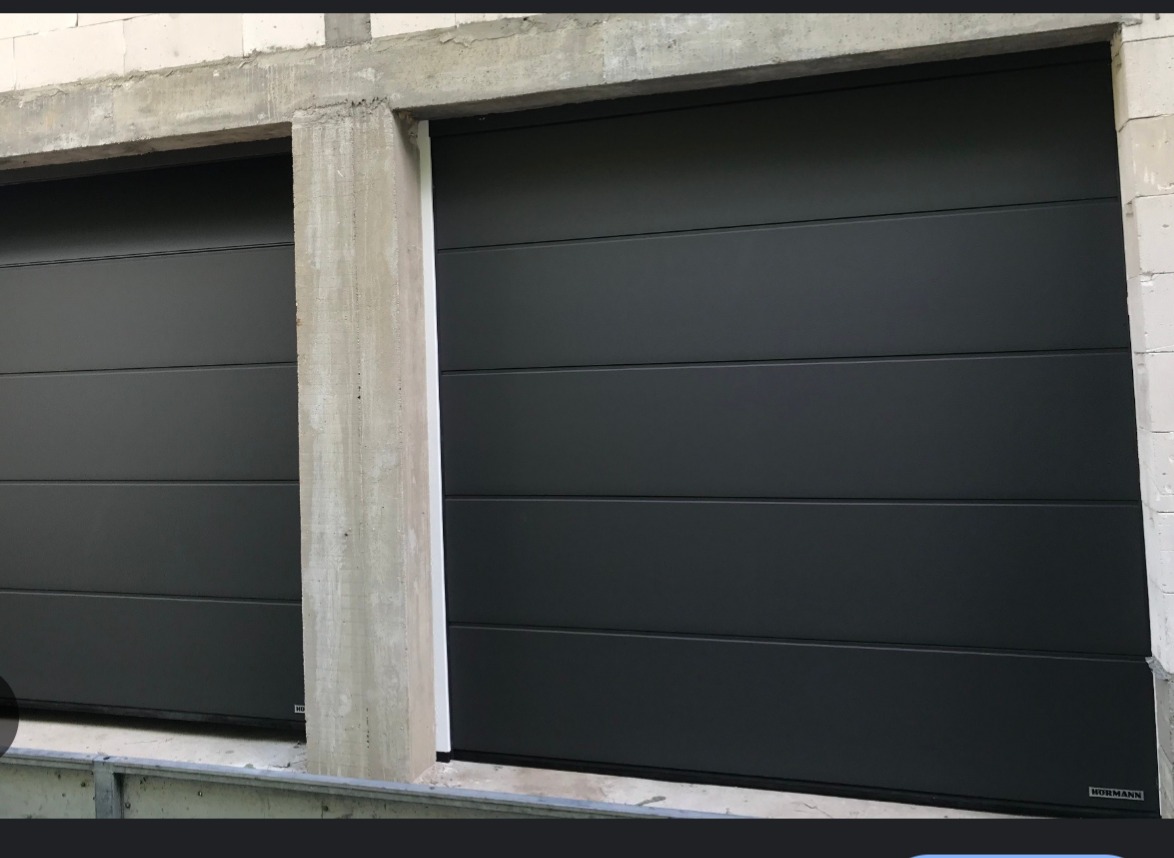 Dwie segmentowe, grafitowe bramy garażowe Hormann w surowym otoczeniu betonowych ścian i nadproży, widoczne detale montażowe.