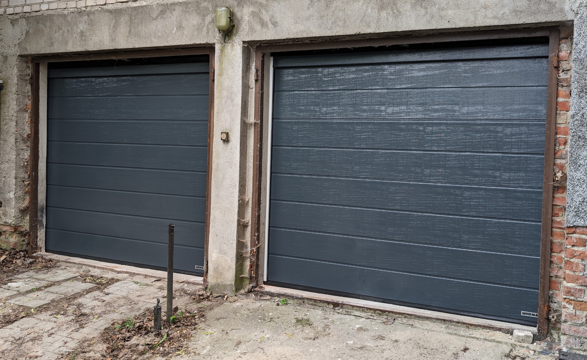 Dwie szare segmentowe bramy garażowe z poziomymi panelami, zamontowane w starym, częściowo otynkowanym budynku z ceglanymi elementami, widoczne ślady użytkowania i zabrudzenia na elewacji.