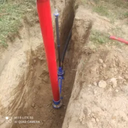 Głęboki wykop w ziemi z widocznymi rurami: czerwoną pionową rurą o dużej średnicy oraz czarną rurą o mniejszej średnicy połączoną z niebieskim zaworem. Na krawędzi wykopu leży poziomica.