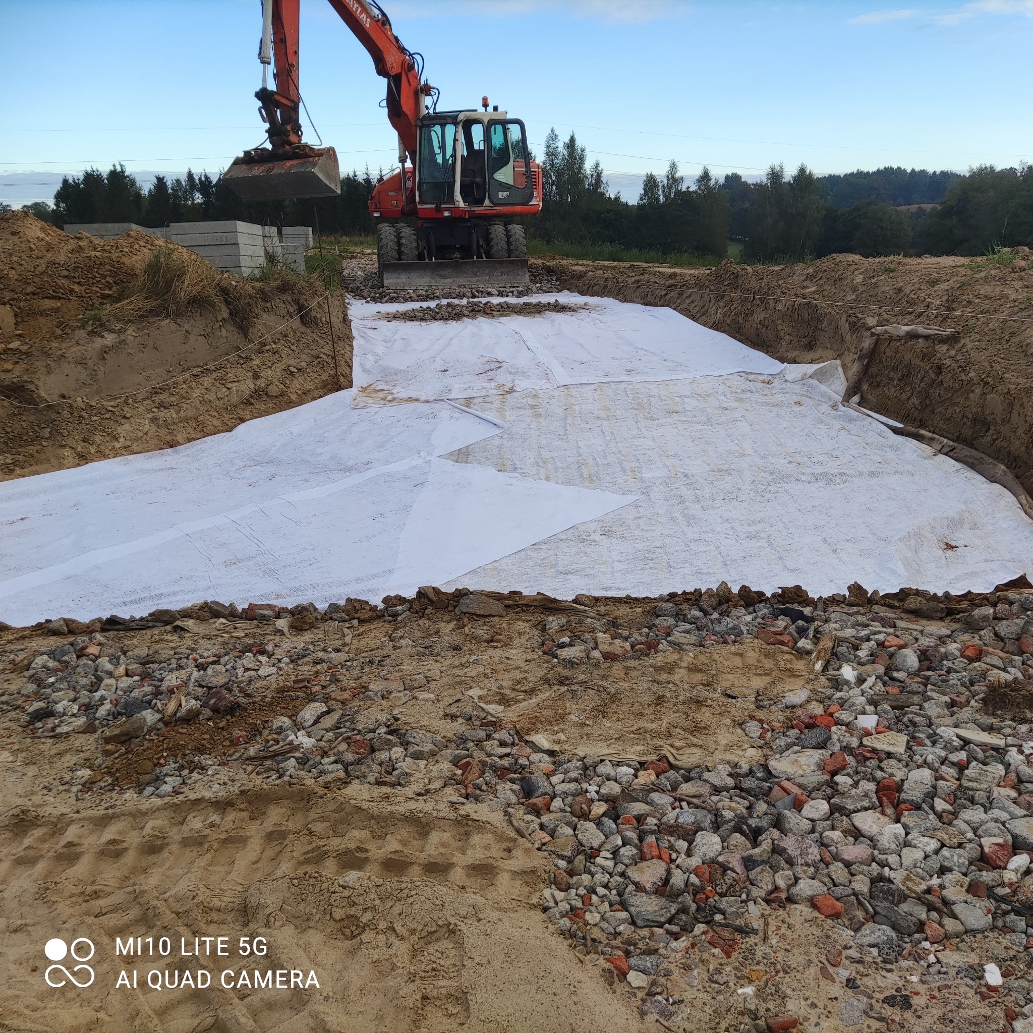 Czerwona koparka Atlas na placu budowy, rozkładanie białej geowłókniny na wyrównanym terenie, widoczne gruz i ziemia, w tle drzewa.