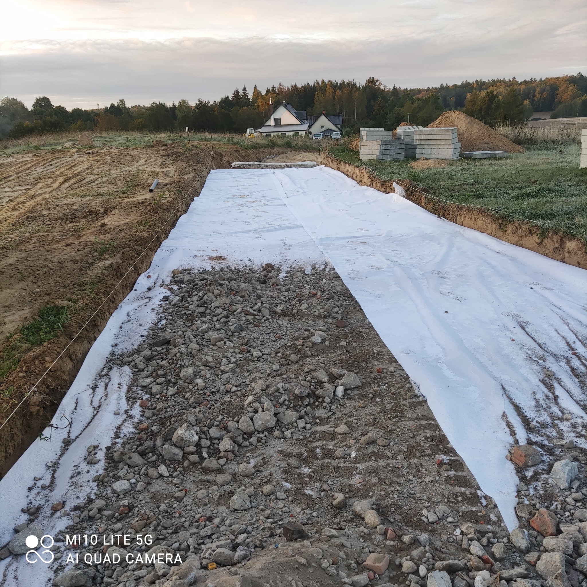 Szeroki wykop zabezpieczony białą geowłókniną, częściowo zasypany kamieniami i gruzem, w tle dom jednorodzinny i las.