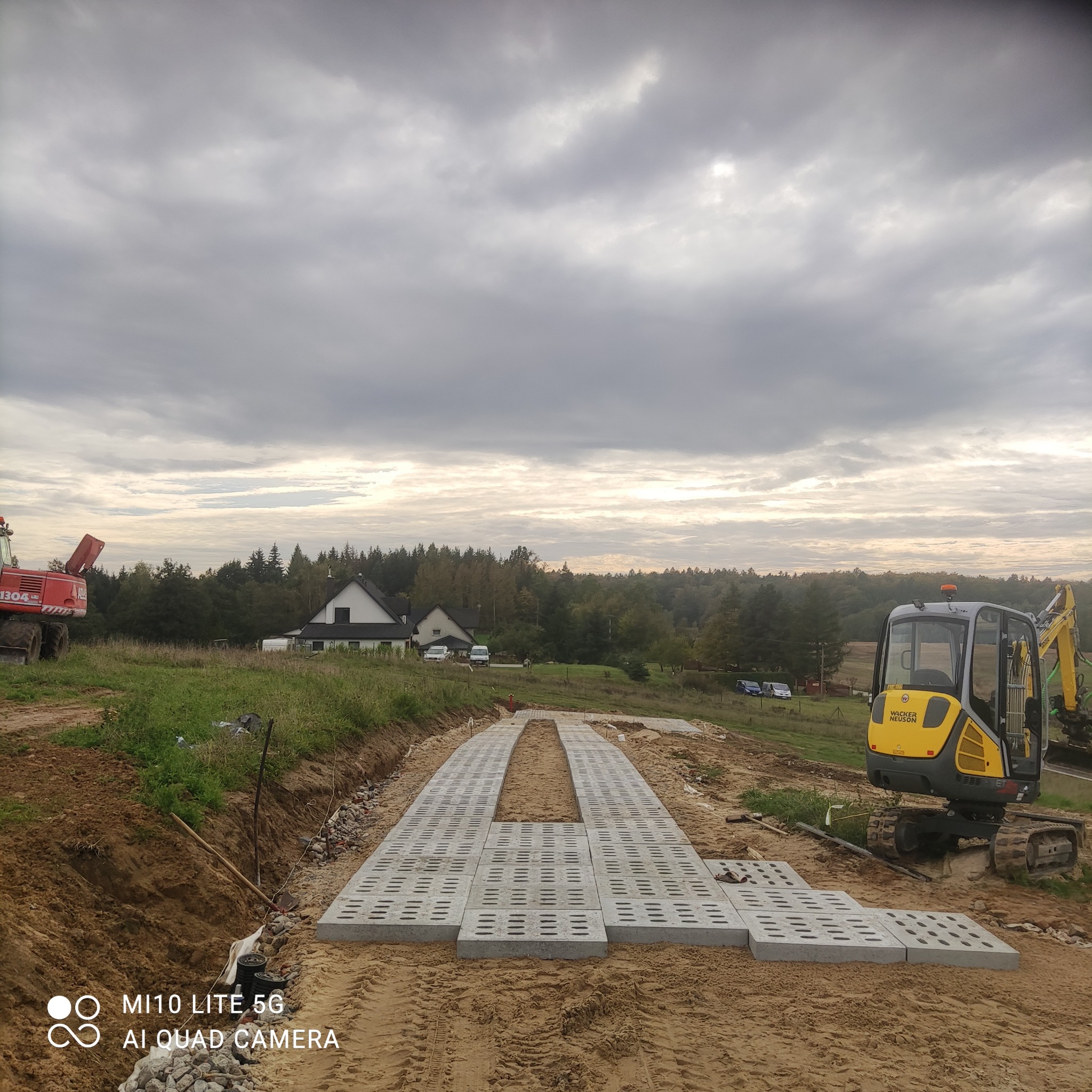 Układanie betonowych płyt ażurowych na piasku, przygotowanie podłoża pod drogę, widoczny mały traktor i koparka Wacker Neuson w tle, pochmurne niebo.