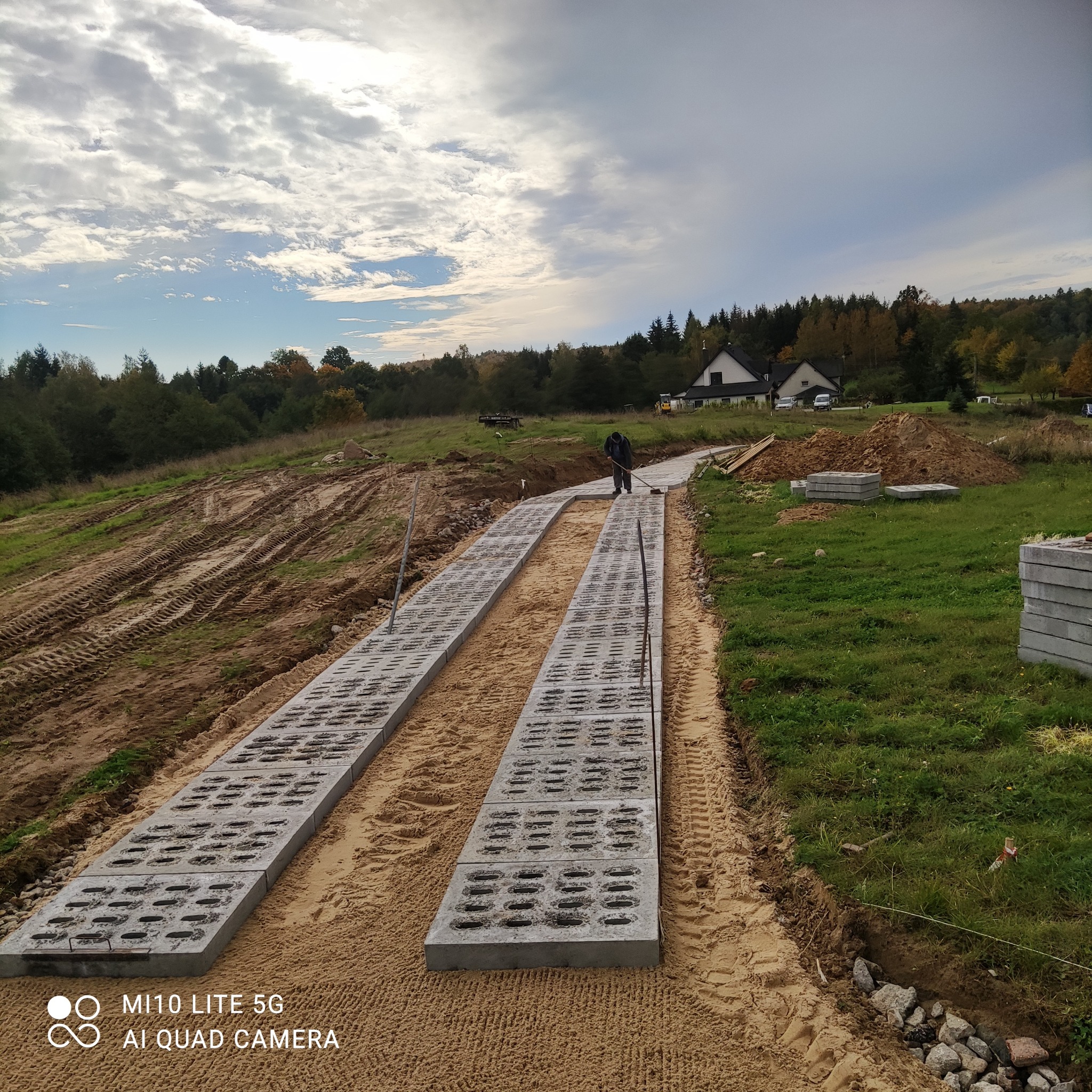 Układanie betonowych płyt ażurowych na piasku w celu wzmocnienia drogi, widoczna osoba wyrównująca powierzchnię za pomocą miotły, w tle dom i drzewa.
