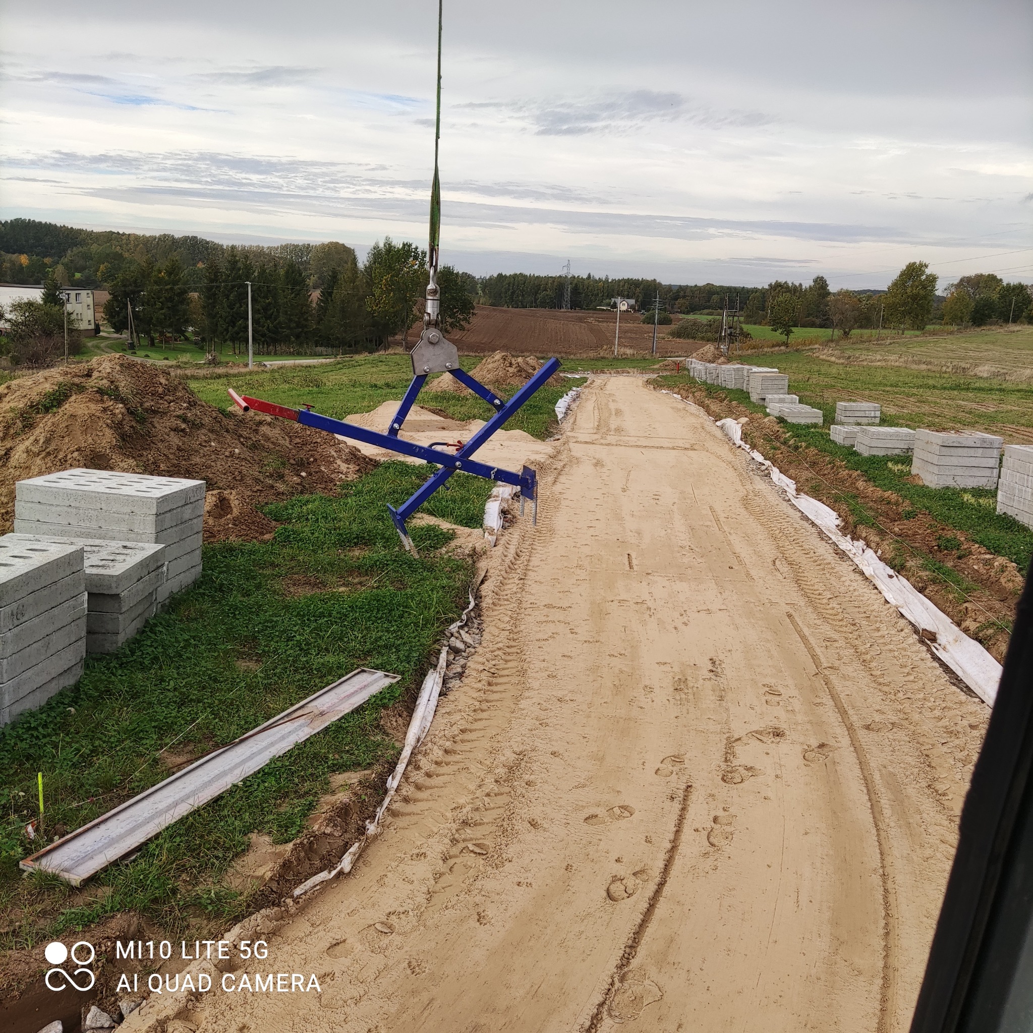 Montaż betonowych elementów koryta drogi za pomocą dźwigu z chwytakiem; widoczne ułożone bloczki, przygotowany teren i ślady opon