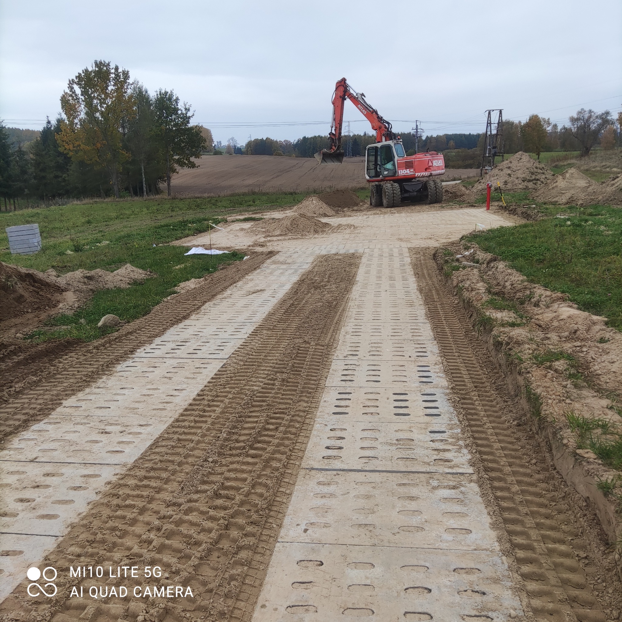 Ciągnik Atlas 1304 w trakcie prac na placu budowy, widoczne płyty drogowe tymczasowe, w tle drzewa i pole uprawne.