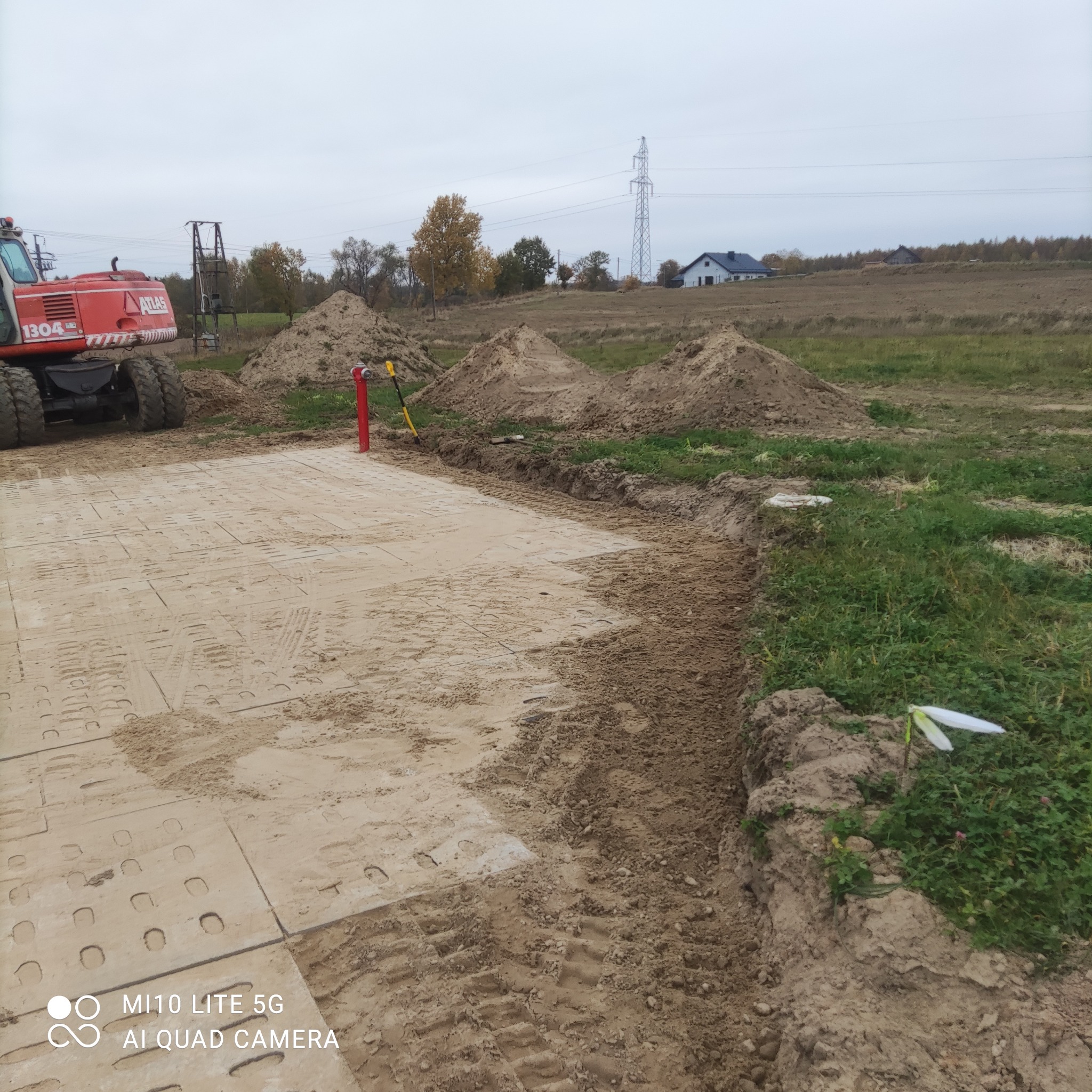 Przygotowany teren z użyciem płyt drogowych, widoczny wykop i hałdy ziemi w tle, na skraju pola, z fragmentem czerwonej koparki kołowej Atlas 1304.