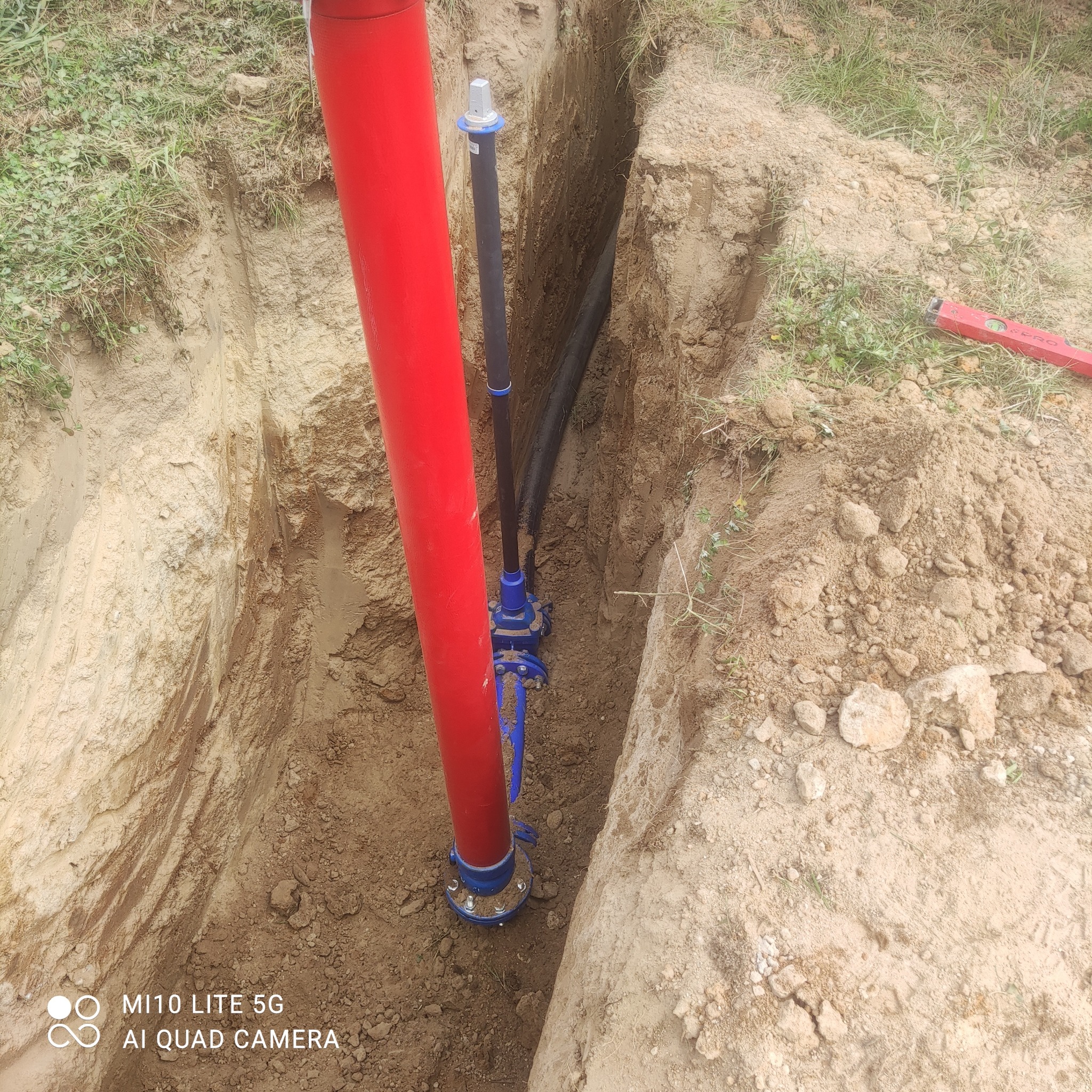 Głęboki wykop w ziemi z widocznymi rurami: czerwoną pionową rurą o dużej średnicy oraz czarną rurą o mniejszej średnicy połączoną z niebieskim zaworem. Na krawędzi wykopu leży poziomica.