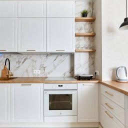 Wysmeble - Biała kuchnia z marmurowym backsplashiem, złotą armaturą i drewnianym blatem; wiszące półki z dekoracjami i czarne lampy.