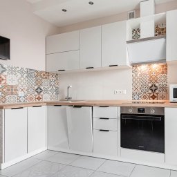 Wysmeble - Biała, narożna zabudowa kuchenna z drewnianym blatem i dekoracyjnymi płytkami w stylu patchwork zaaranżowana w mieszkaniu w Białymstoku. Na ścianie wisi telewizor.