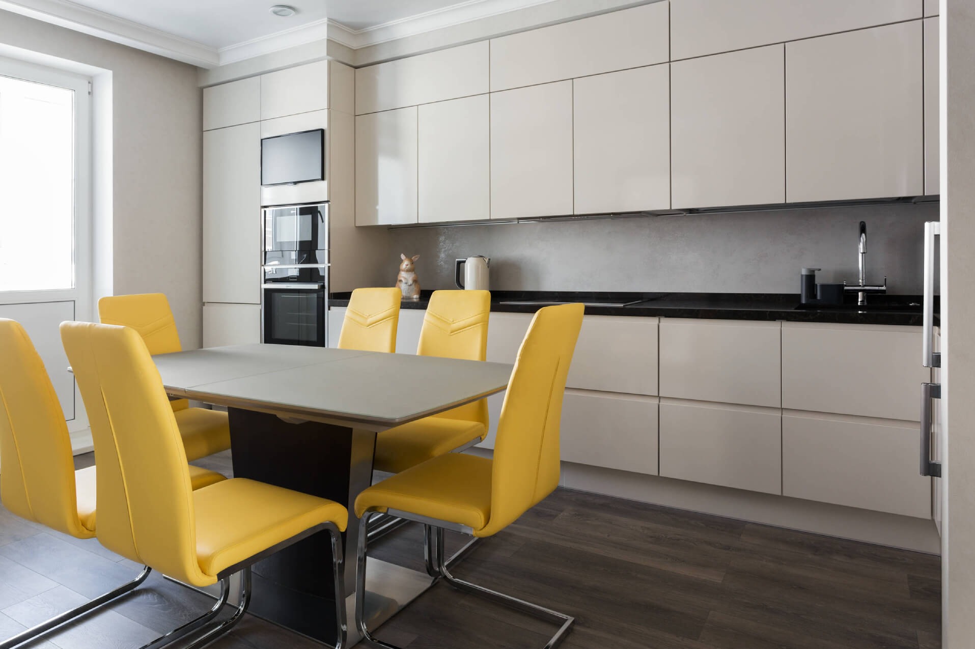 Jasna kuchnia z zabudową na wymiar, czarnym blatem i żółtymi krzesłami przy stole. W tle widoczny sprzęt AGD w zabudowie i dekoracyjny królik.