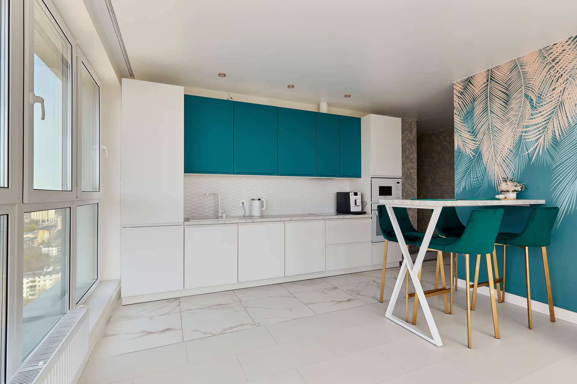 Nowoczesna, jasna kuchnia z białymi szafkami dolnymi, turkusowymi szafkami górnymi i blatem imitującym marmur, uzupełniona o wysoki stół z białym blatem i zielonymi krzesłami barowymi na złotych...