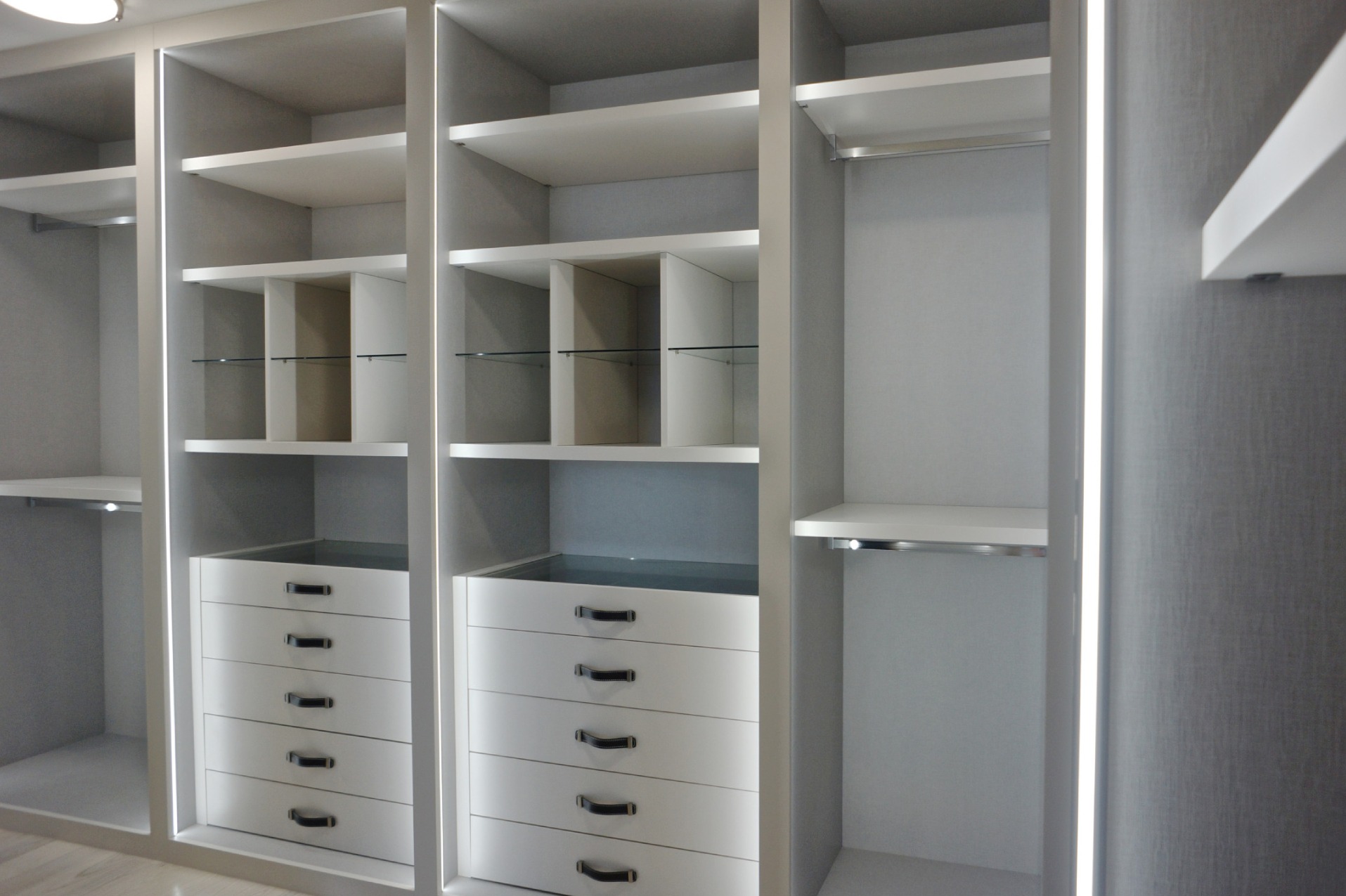 Wnętrze szafy wnękowej z szufladami, półkami i drążkiem na ubrania, oświetlenie LED w pionowych krawędziach, jasna kolorystyka.