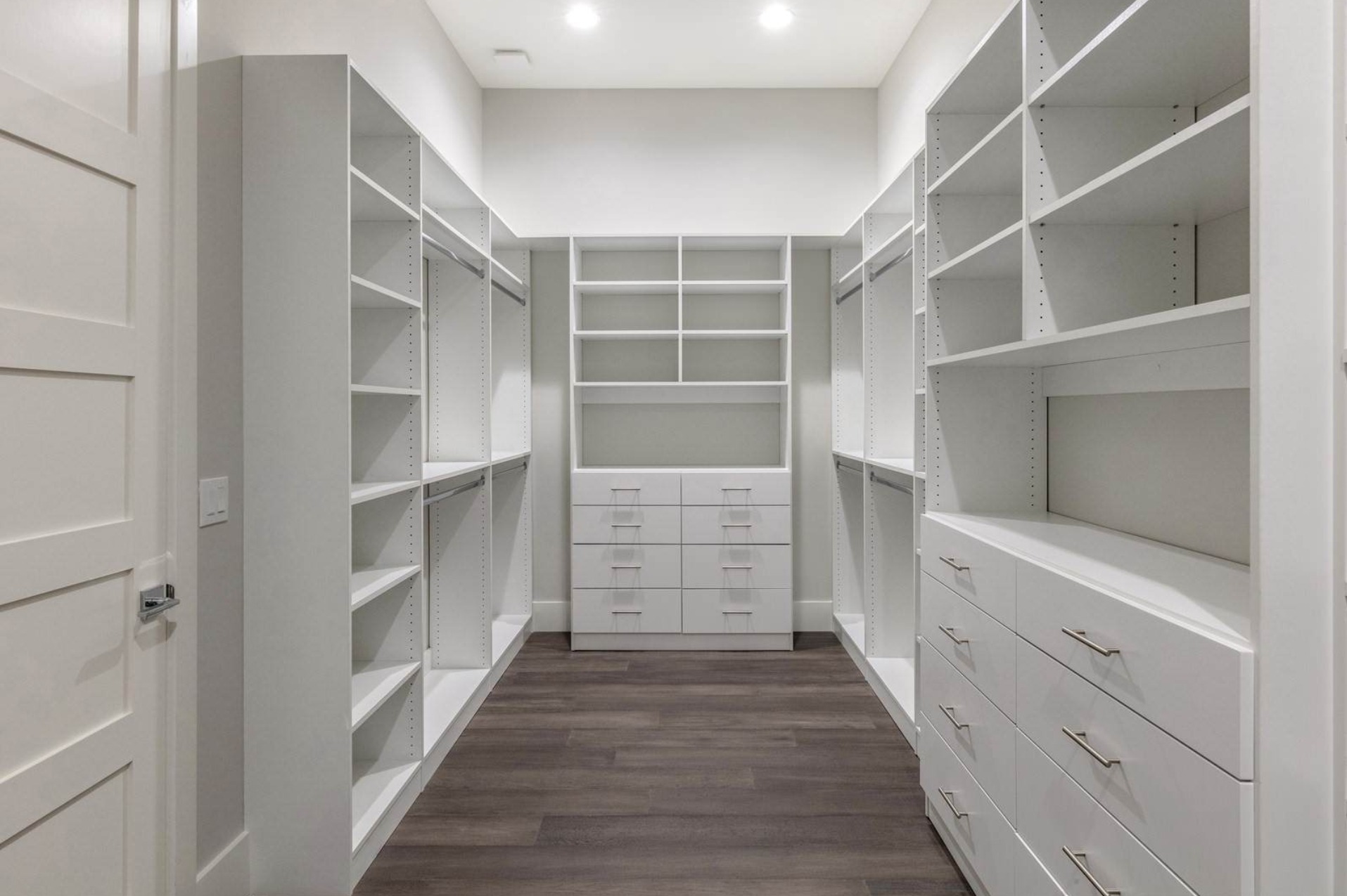 Biała, przestronna garderoba z półkami i szufladami, widok z perspektywy wejścia, ciemna podłoga.