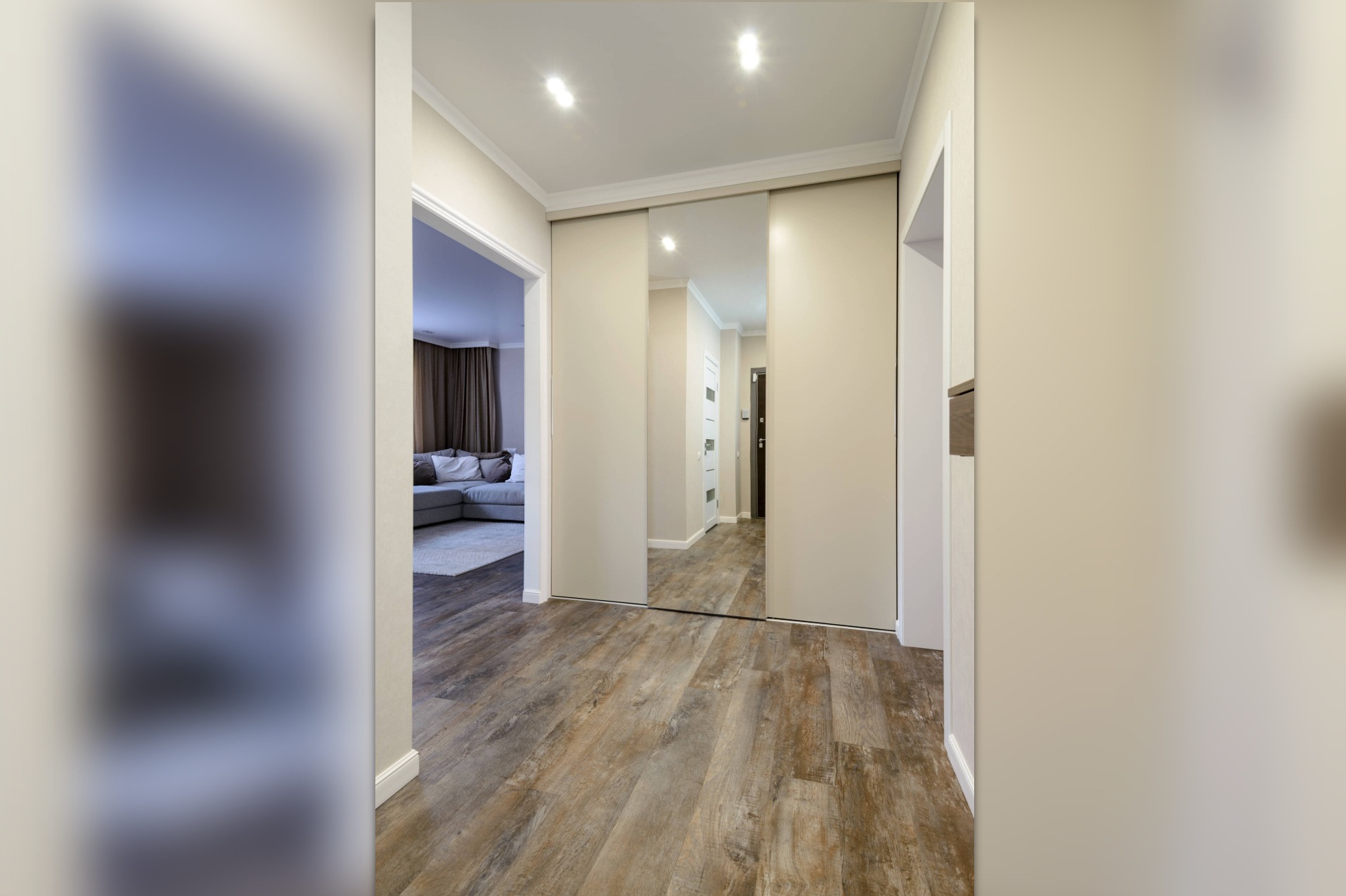 Beżowa szafa przesuwna z lustrem w przedpokoju, widoczna drewniana podłoga i fragment salonu z sofą.