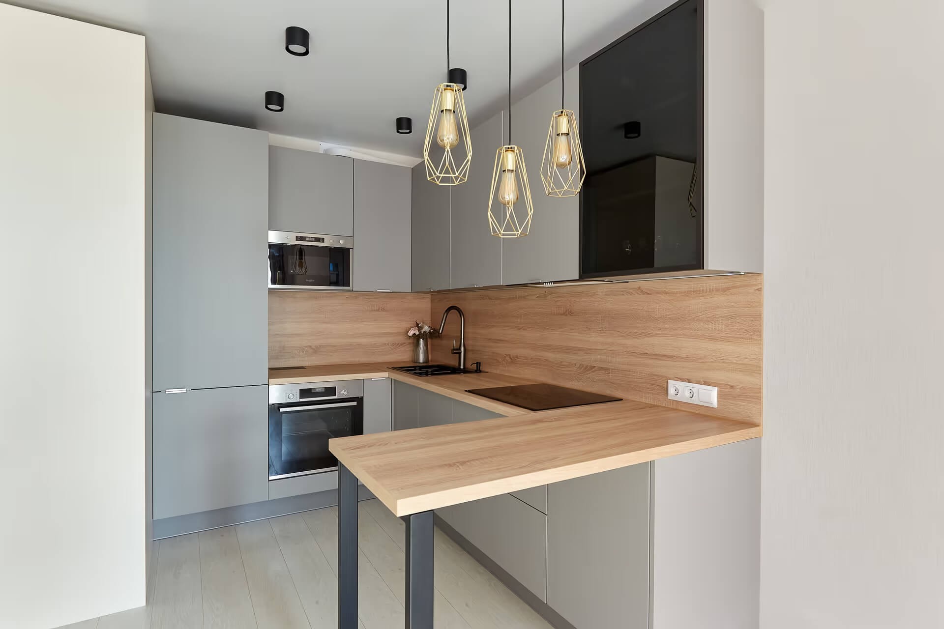 Nowoczesna kuchnia w odcieniach szarości z drewnianym blatem i złotymi lampami wiszącymi o geometrycznym kształcie, widok na zabudowę kuchenną z piekarnikiem i mikrofalówką.