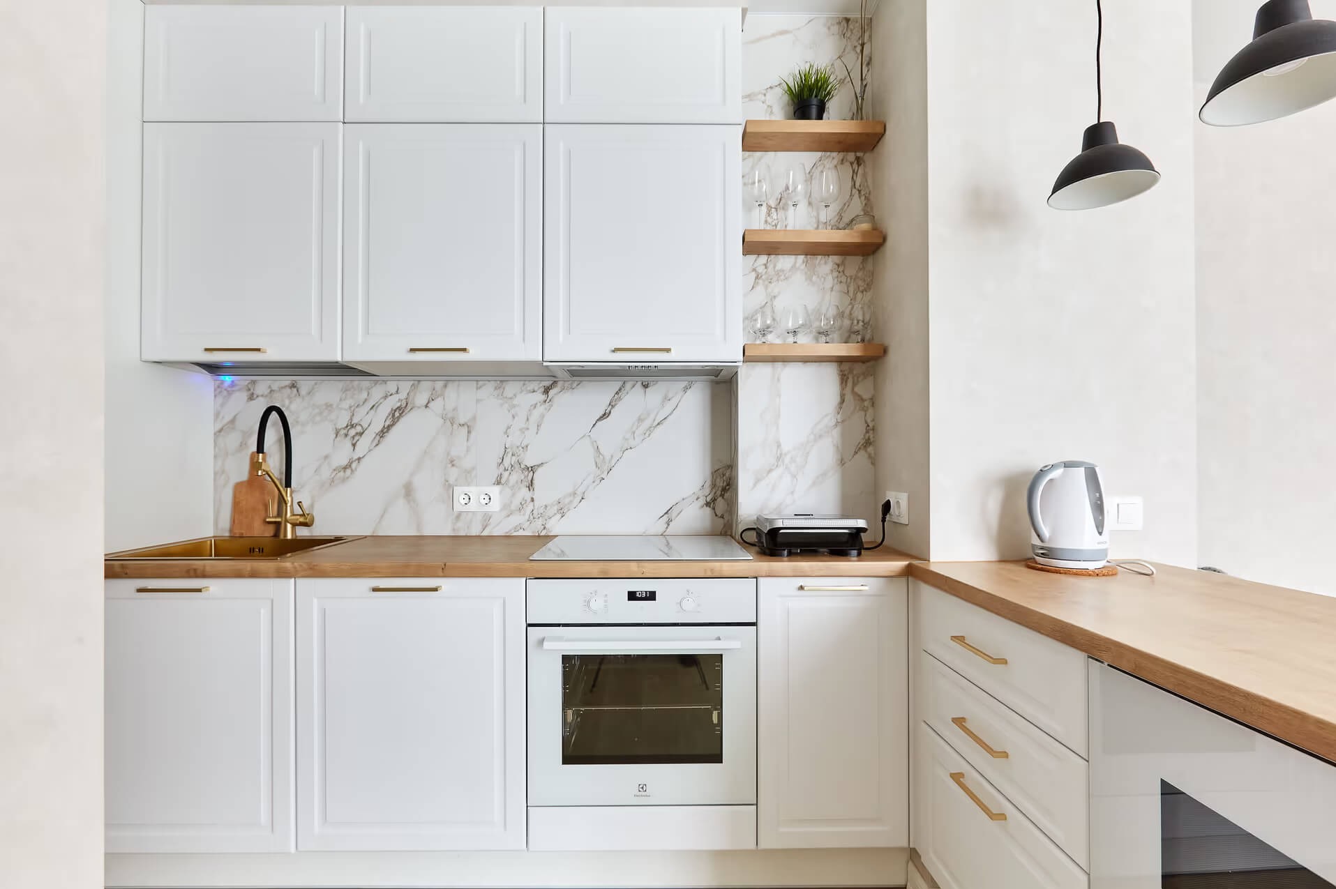 Biała kuchnia z marmurowym backsplashiem, złotą armaturą i drewnianym blatem; wiszące półki z dekoracjami i czarne lampy.