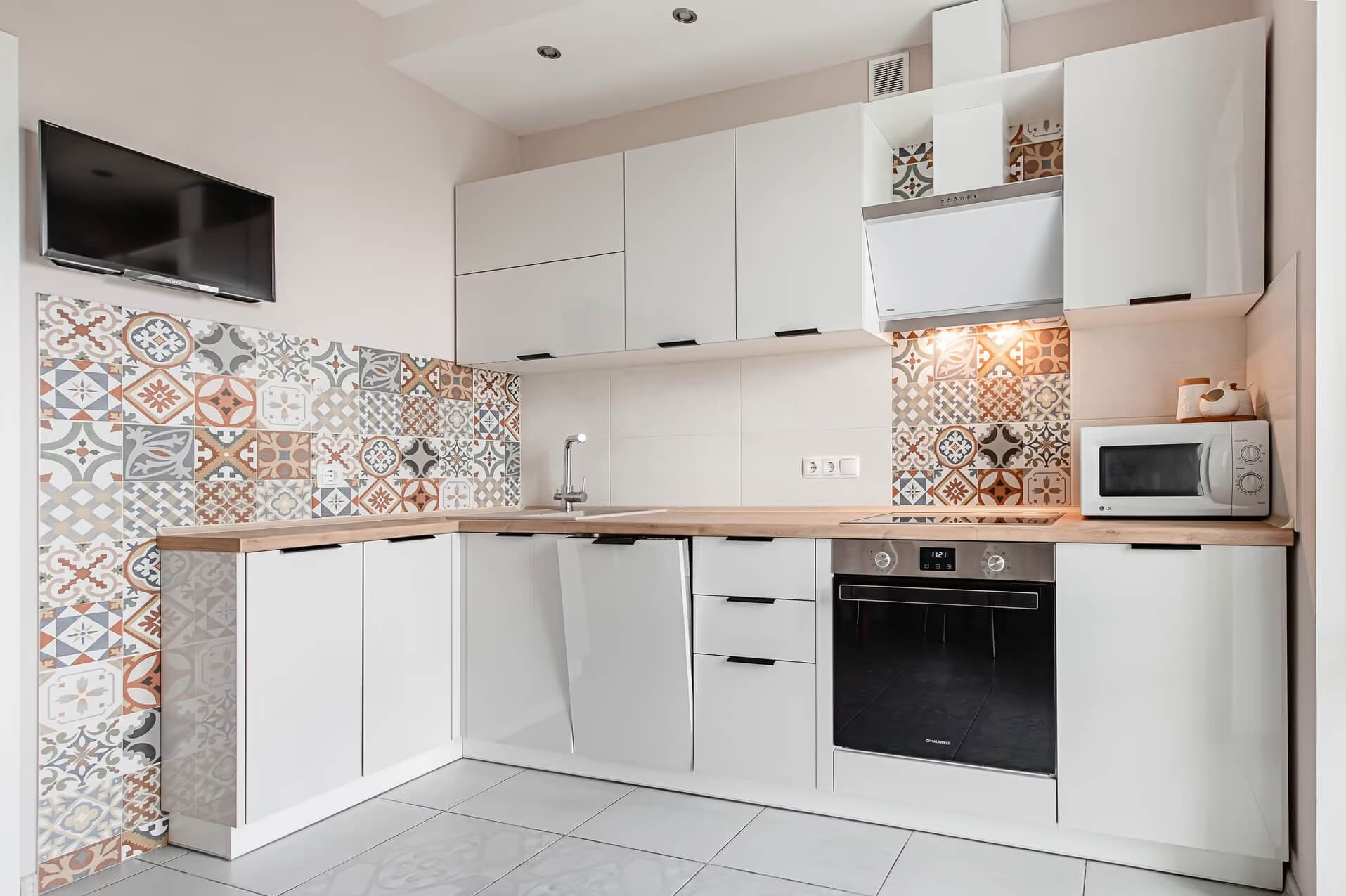 Biała, narożna zabudowa kuchenna z drewnianym blatem i dekoracyjnymi płytkami w stylu patchwork zaaranżowana w mieszkaniu w Białymstoku. Na ścianie wisi telewizor.