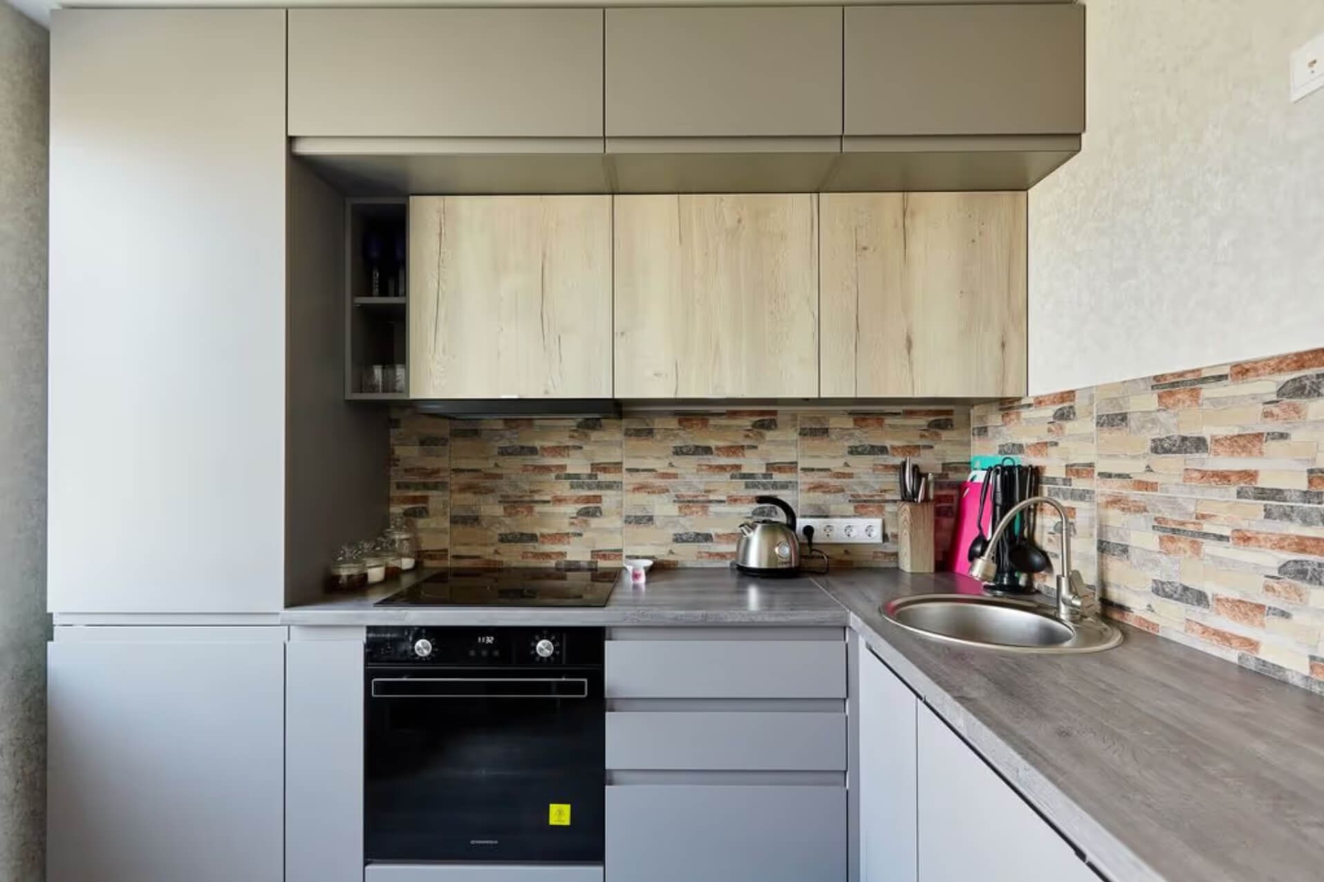 Szara kuchnia na wymiar z górnymi szafkami w kolorze drewna, z płytkami imitującymi cegłę na ścianie i czarnym piekarnikiem.