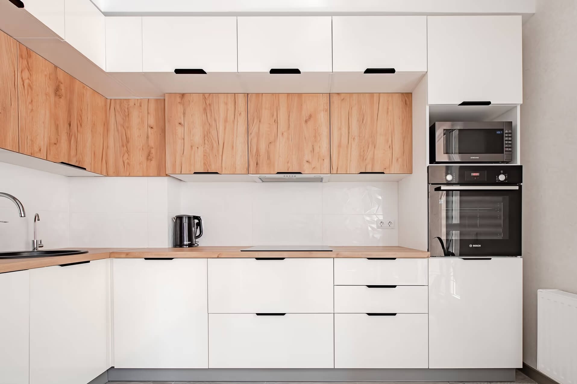 Nowoczesna kuchnia z białymi frontami dolnych szafek i drewnopodobnymi górnymi szafkami, czarne, minimalistyczne uchwyty, zabudowany piekarnik i mikrofalówka.
